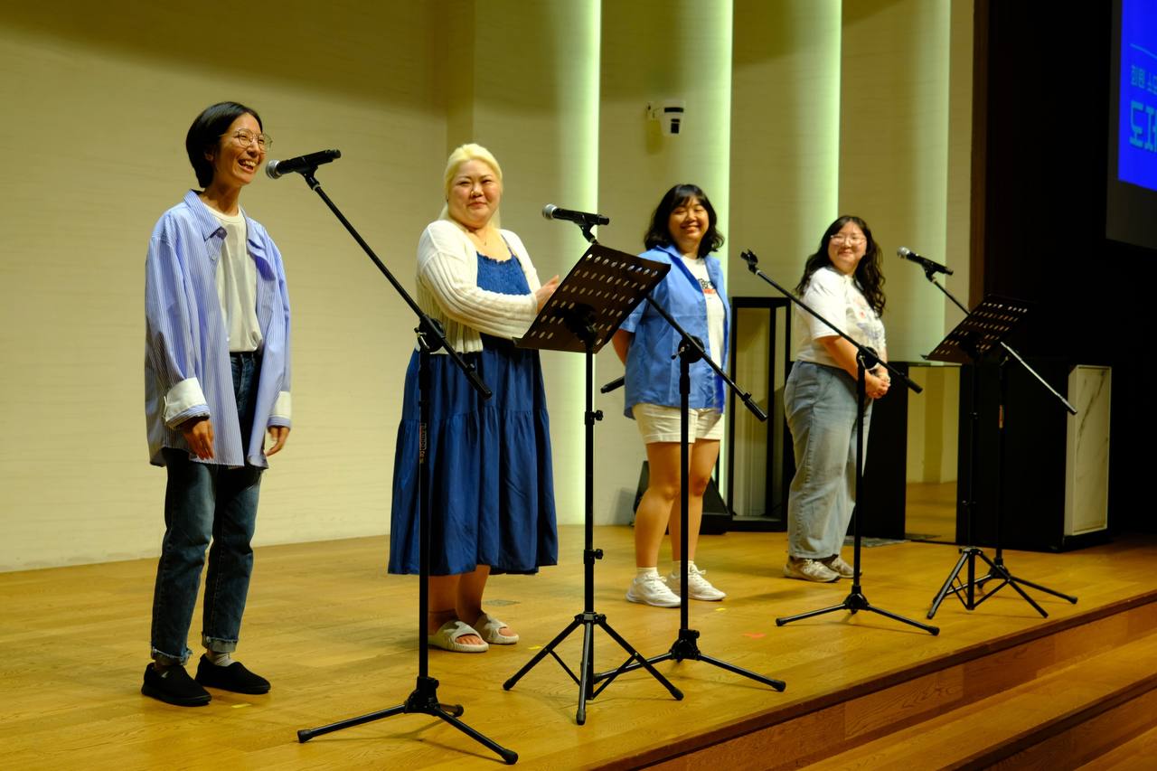 "무대 위에 네 명의 여성이 나란히 서서 관객을 향해 미소 짓고 있다. 각각 마이크 앞에 서 있으며, 왼쪽부터 청바지와 줄무늬 셔츠, 파란색 원피스, 파란 셔츠와 흰 반바지, 청바지와 흰 티셔츠를 입었다. 모두 편안한 분위기에서 대화를 나누듯 즐겁게 서 있다."