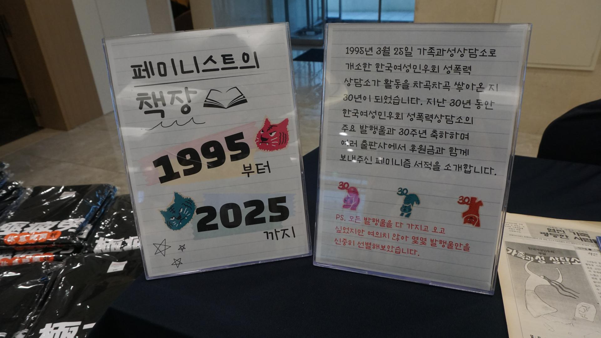 "행사 안내 테이블 위에 두 개의 세워진 안내판이 나란히 놓여 있다. 왼쪽 안내판에는 ‘페미니스트의 책장 1995부터 2025까지’라는 글씨와 함께 호랑이 얼굴 그림과 별 그림이 장식되어 있다. 오른쪽 안내판에는 한국여성민우회 성폭력상담소 30주년을 기념하는 안내문이 적혀 있으며, '1995년 3월 25일 개소한 한국여성민우회 성폭력상담소가 30년이 되었습니다'라는 문장으로 시작한다. 안내판 아래쪽에는 'PS. 모든 발행물을 다 가지고 오고 싶었지만 여의치 않아 몇몇 발행물만을 신중히 선별해보았습니다'라는 문구가 빨간색으로 강조되어 있다. 왼쪽 테이블에는 검은색 티셔츠가 여러 장 포개져 있다."