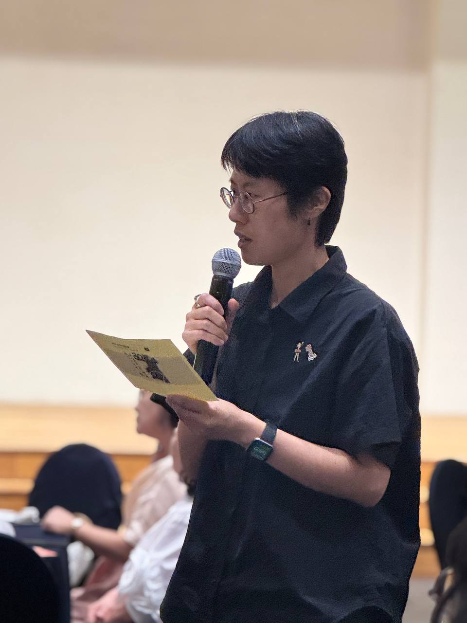 청중석에서 일어나 발표하는 회원 혜영의 모습. 검은 셔츠를 입고 검은색 짧은 머리를 하고 있다. 마이크를 들고 서 있으며 주변엔 앉아 있는 다른 청중들의 모습이 흐릿하게 보인다. 