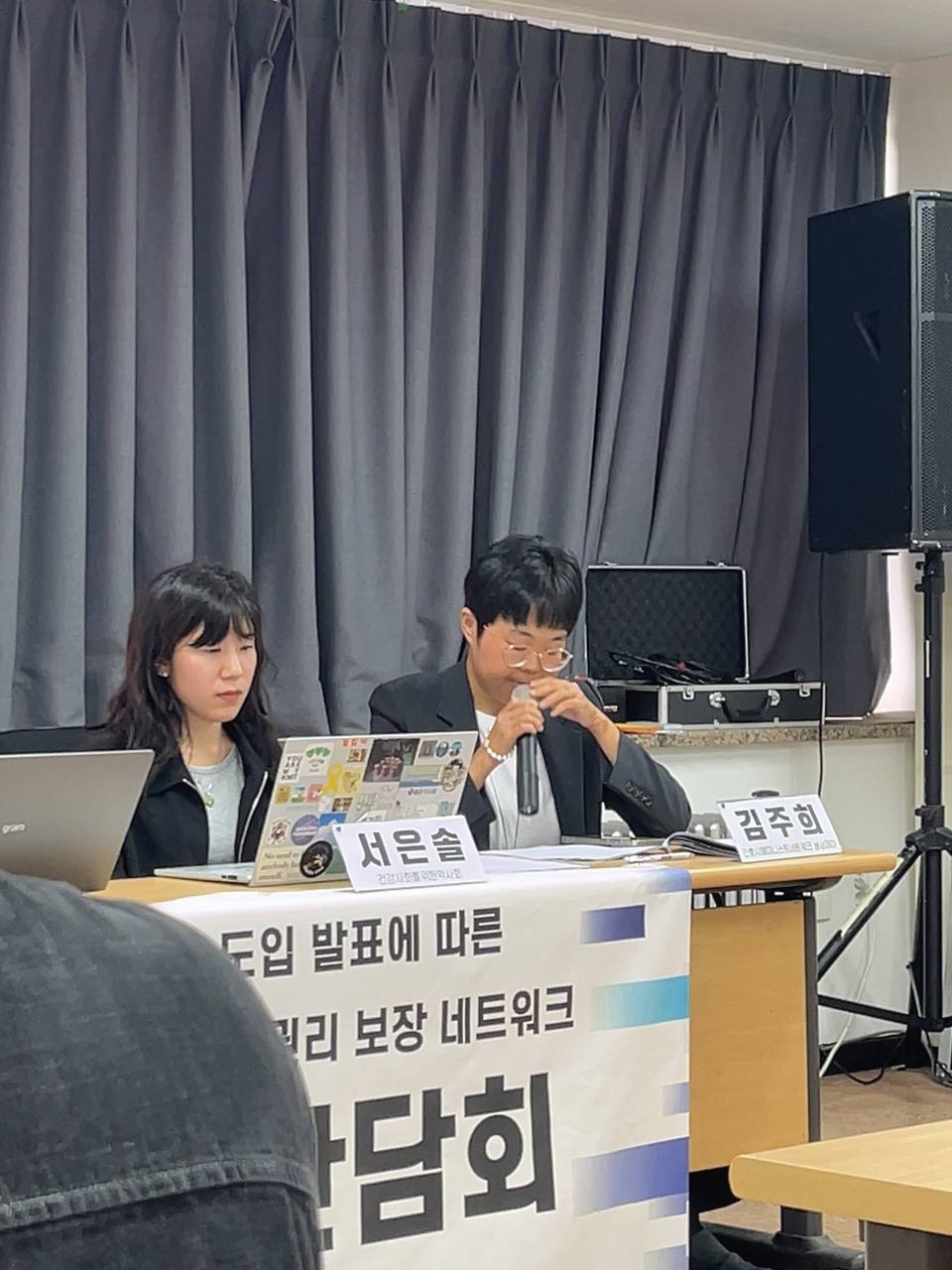 한 사람이 마이크를 들고 발언 중이다. 그 옆으로는 네 명의 사람이 앉아서 자료를 보고 있고, 뒤로는 기자간담회 제목이 적힌 텔레비전이, 책상 앞쪽에는 기자간담회 제목이 적힌 현수막이 걸려 있다.