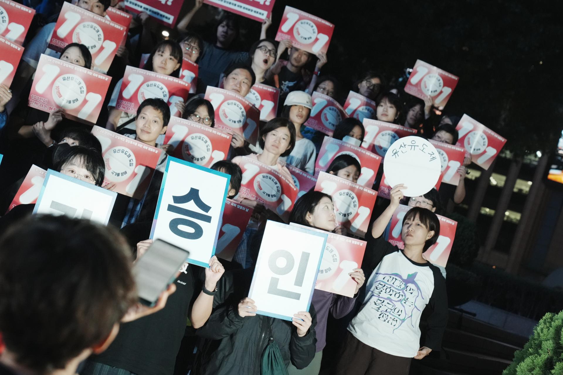 유산유도제라고 적힌 알약 모양의 피켓을 든 민우회 활동가가 사회자를 바라보고 있다. 다른 참여자들은 각자 피켓을 들고 유산유도제 알약 모형을 바라보며 그것이 건네질 순간을 기다리는 중이다. 사진 앞쪽으로는 몽실(사회자)의 뒷모습이 보인다.