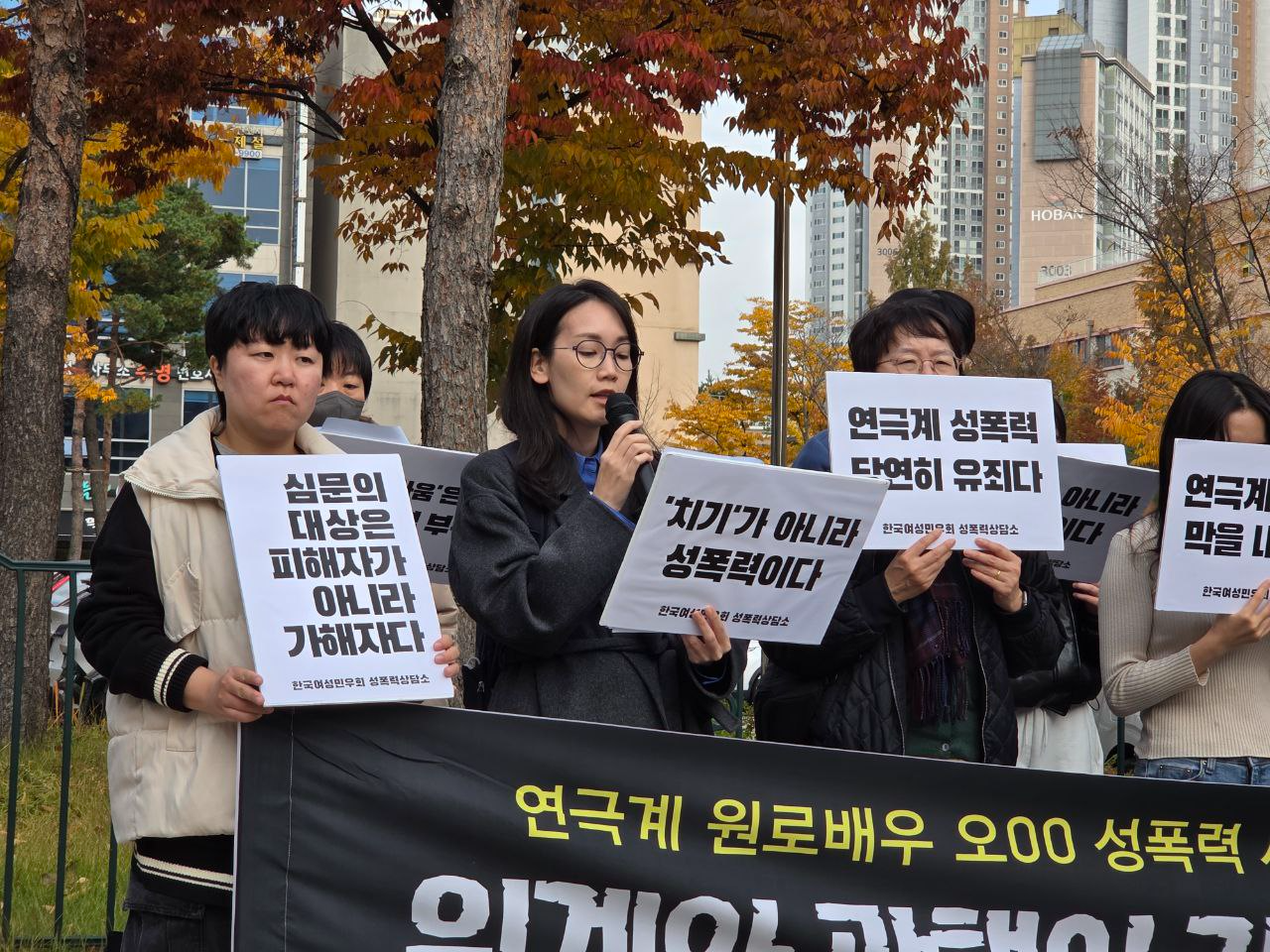 사람들이 연극계 원로배우 오00 성폭력 사건 항소심 판결에 분노하는 문구가 담긴 각각의 피켓을 들고 침통한 표정으로 서있다. 가운데 안경을 쓴 단발머리 여성이 마이크를 들고 발언하고 있다.