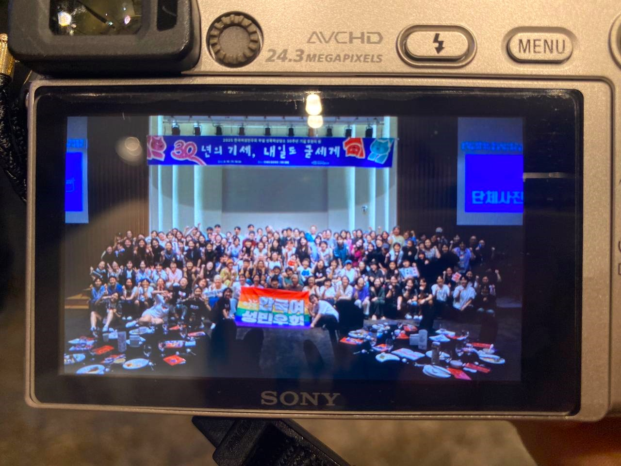 "소니 디지털카메라 미리보기 화면의 모습. 실내 행사장에서 150여 명이 무대 위에 모여 단체사진을 찍고 있다. 가운데에 무지개 색 배너를 든 사람들이 있고, 배너에는 '한국여성민우회'라는 글자가 적혀 있다. 무대 뒤에는 파란색 현수막이 걸려 있으며, ‘30년의 기세, 내일도 굳세게’라는 문구와 함께 빨간색과 파란색 그림이 보인다. 무대 앞쪽 테이블 위에는 식사 접시와 음료가 놓여 있다."