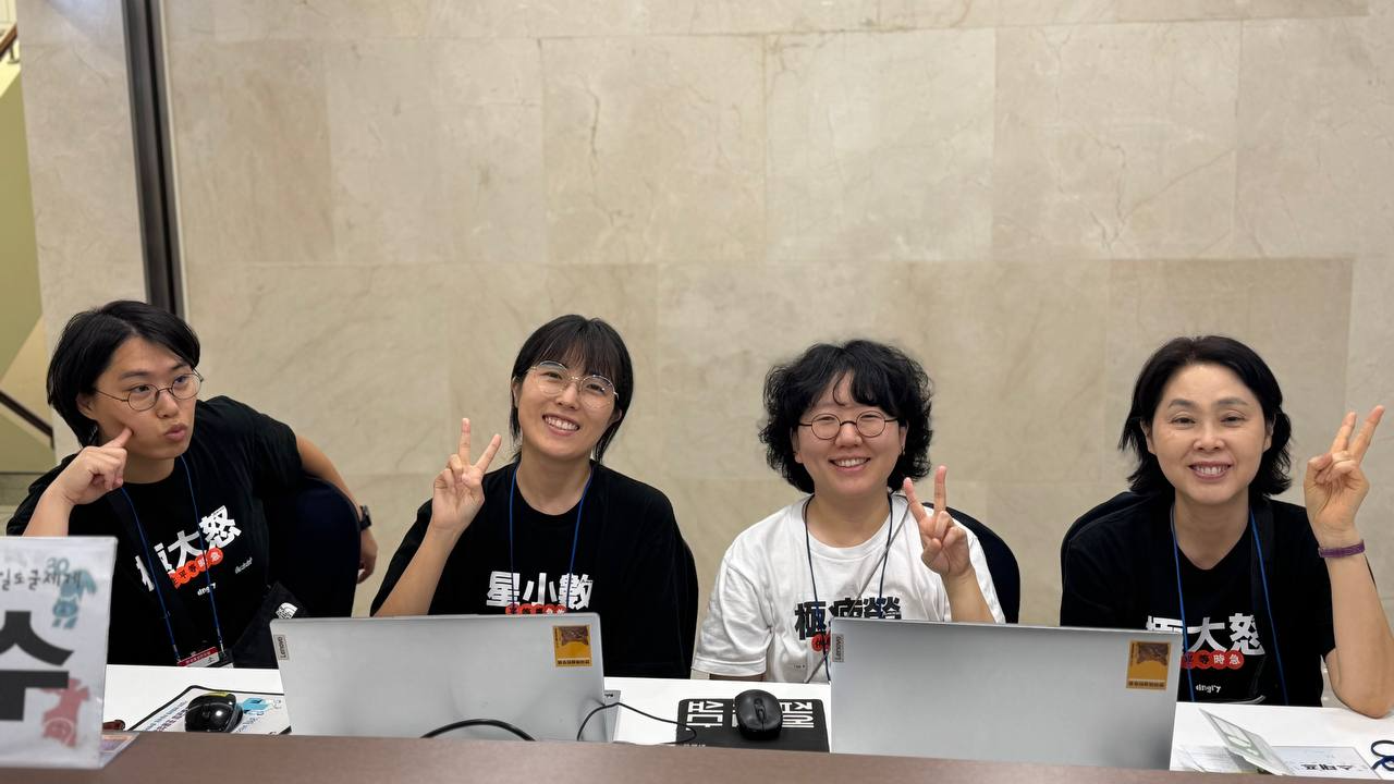 접수대 앞에 앉은 네 명의 활동가들이 웃으며 포즈를 취하고 카메라를 보고 있는 모습 