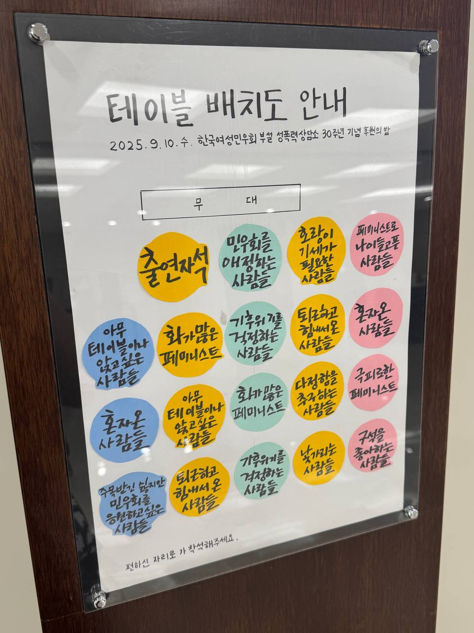 "갈색 판에 붙은 ‘테이블 배치도 안내’ 포스터. 상단에는 ‘2025년 9월 10일, 한국여성민우회 부설 성폭력상담소 30주년 기념 후원의 밤’이라는 글씨가 적혀 있다. 포스터 중앙에는 사각형으로 ‘무대’라고 표시되어 있고, 그 아래에 여러 개의 둥근 색깔 원이 배치되어 테이블을 나타낸다. 각 원 안에는 ‘출연석’, ‘민우회를 애정하는 사람들’, ‘화가 많은 페미니스트’, ‘기후위기를 걱정하는 사람들’, ‘퇴근하고 힘내서 온 사람들’, ‘주목받고 싶지만 민우회를 응원하고 싶은 사람들’, ‘혼자인 사람들’, ‘낯가리는 사람들’, ‘규칙을 좋아하는 사람들’ 등 다양한 유머러스한 문구가 적혀 있어 사람들이 편한 자리에 앉을 수 있도록 안내하고 있다."