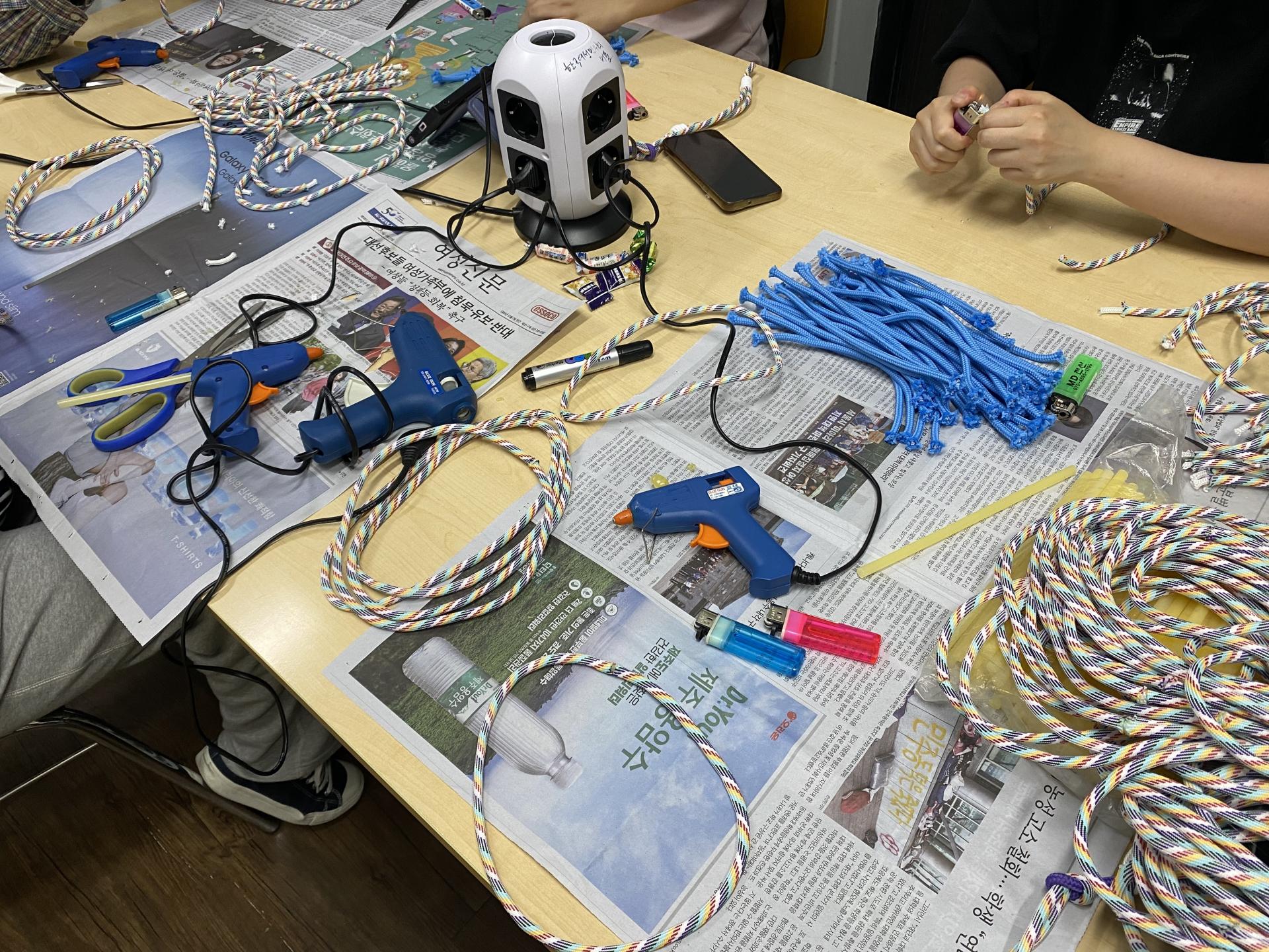 휴대폰 스트랩을 만들고 있는 사진. 책상 위에 글루건, 라이터, 스트랩 로프가 올려져 있다.