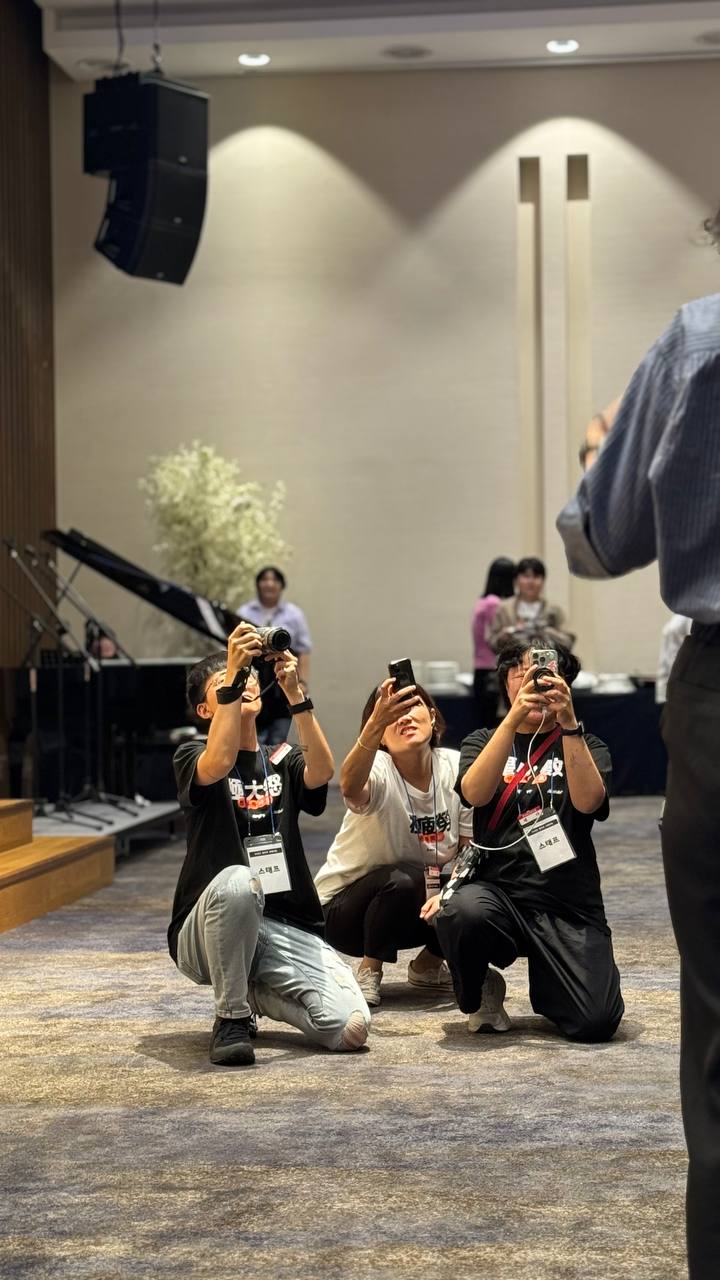 스탭 명찰을 목에 건 활동가 세 명이 바닥에 무릎을 꿇고 앉아 각자의 카메라와 핸드폰카메라를 얼굴 높이 위로 들고, 그 앞에 서서 말하는 누군가를 촬영하고 있는 모습.