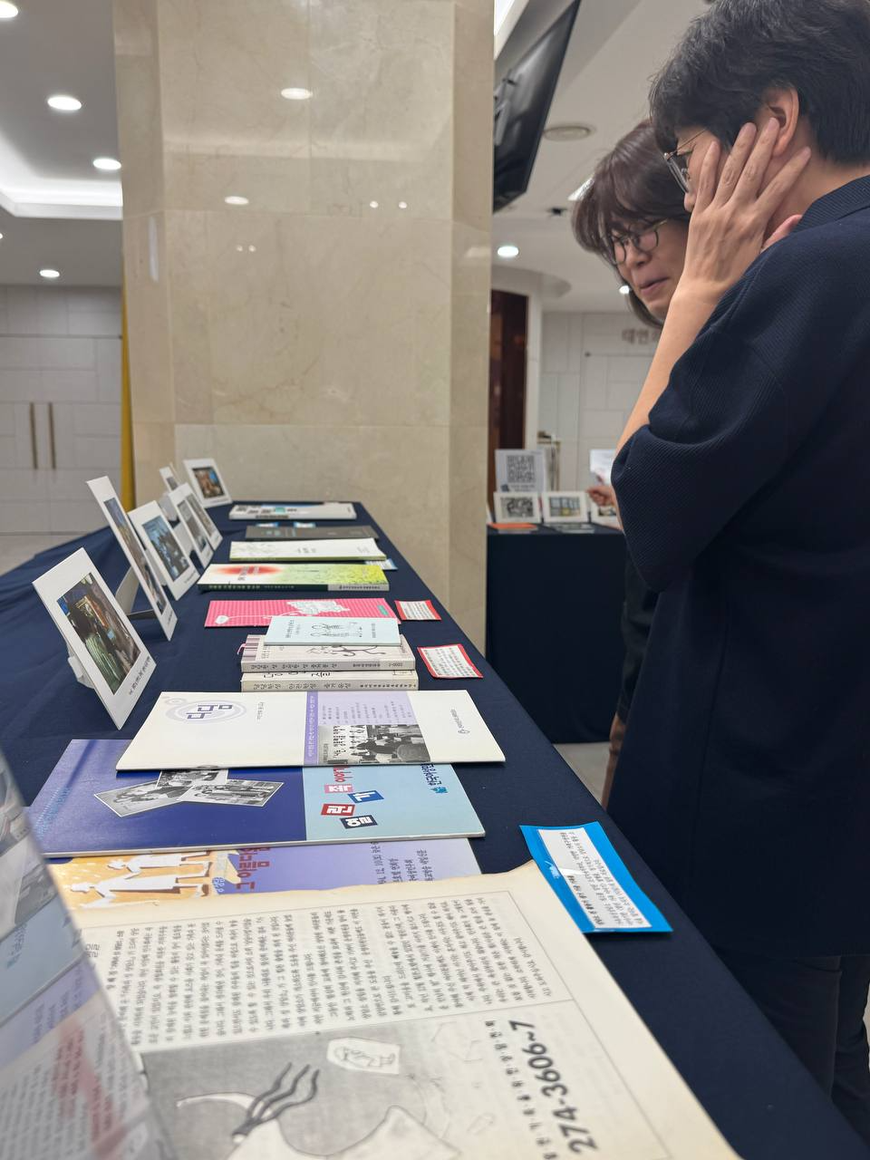 두 명의 사람이 테이블 앞에 서서 테이블 위에 전시된 책자와 사진을 보고 있는 모습.