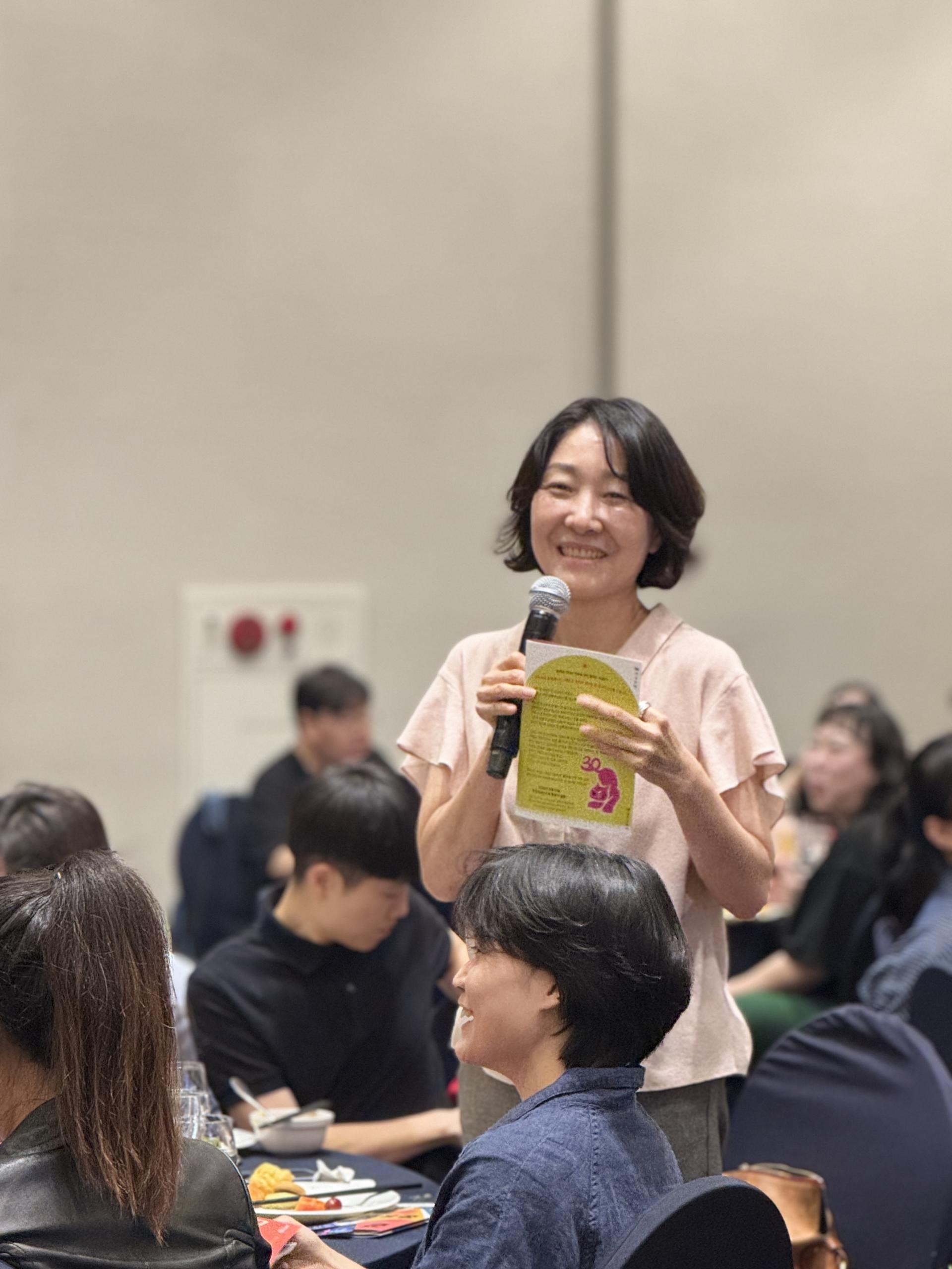 여러 사람이 앉아있는 가운데 분홍색 셔츠를 입은 한 사람이 마이크를 들고 웃으며 일어나 이야기하는 모습