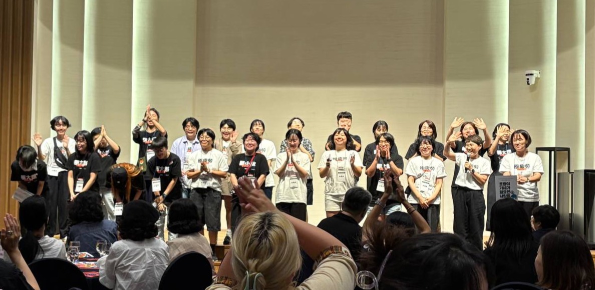 "무대 위에 20여 명의 사람들이 줄지어 서서 인사하고 있다. 흰색과 검은색 티셔츠를 입은 이들이 손을 흔들거나 박수치며 웃고 있고, 객석에서는 사람들이 박수를 보내고 있다."