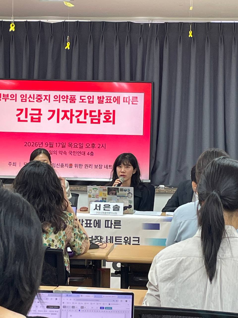 한 사람이 마이크를 들고 발언 중이다. 그 옆으로는 네 명의 사람이 앉아서 자료를 보고 있고, 뒤로는 기자간담회 제목이 적힌 텔레비전이, 책상 앞쪽에는 기자간담회 제목이 적힌 현수막이 걸려 있다.