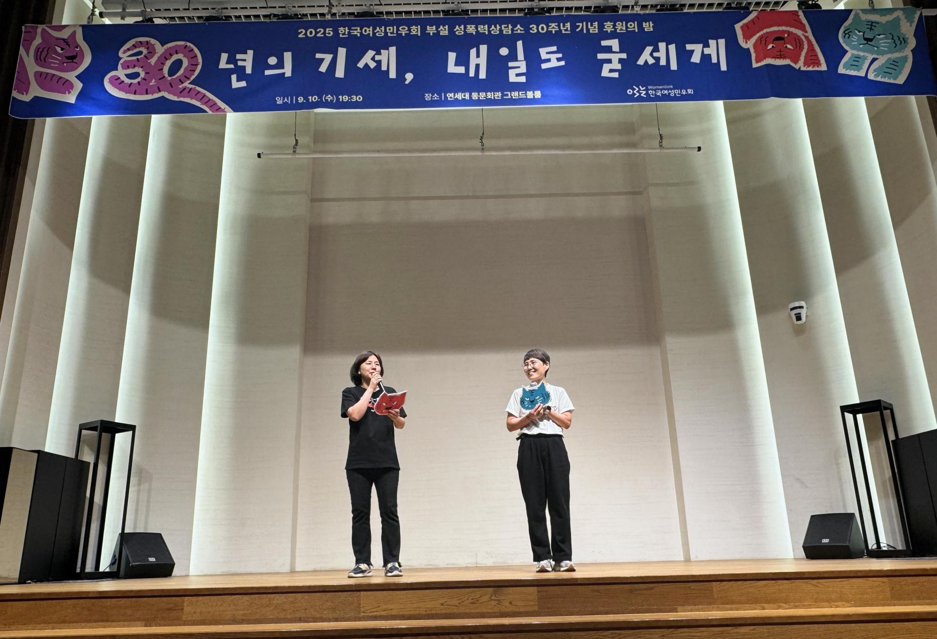 무대 중앙에 두 사람이 서 있다. 둘 다 각각 빨간색, 파란색 호랑이 얼굴 모양 피켓을 손에 들고 있고, 한 명이 마이크를 잡고 말하고 있다. 무대 위쪽에 푸른색 현수막에 후원의밤 행사 제목이 적혀 있다. 