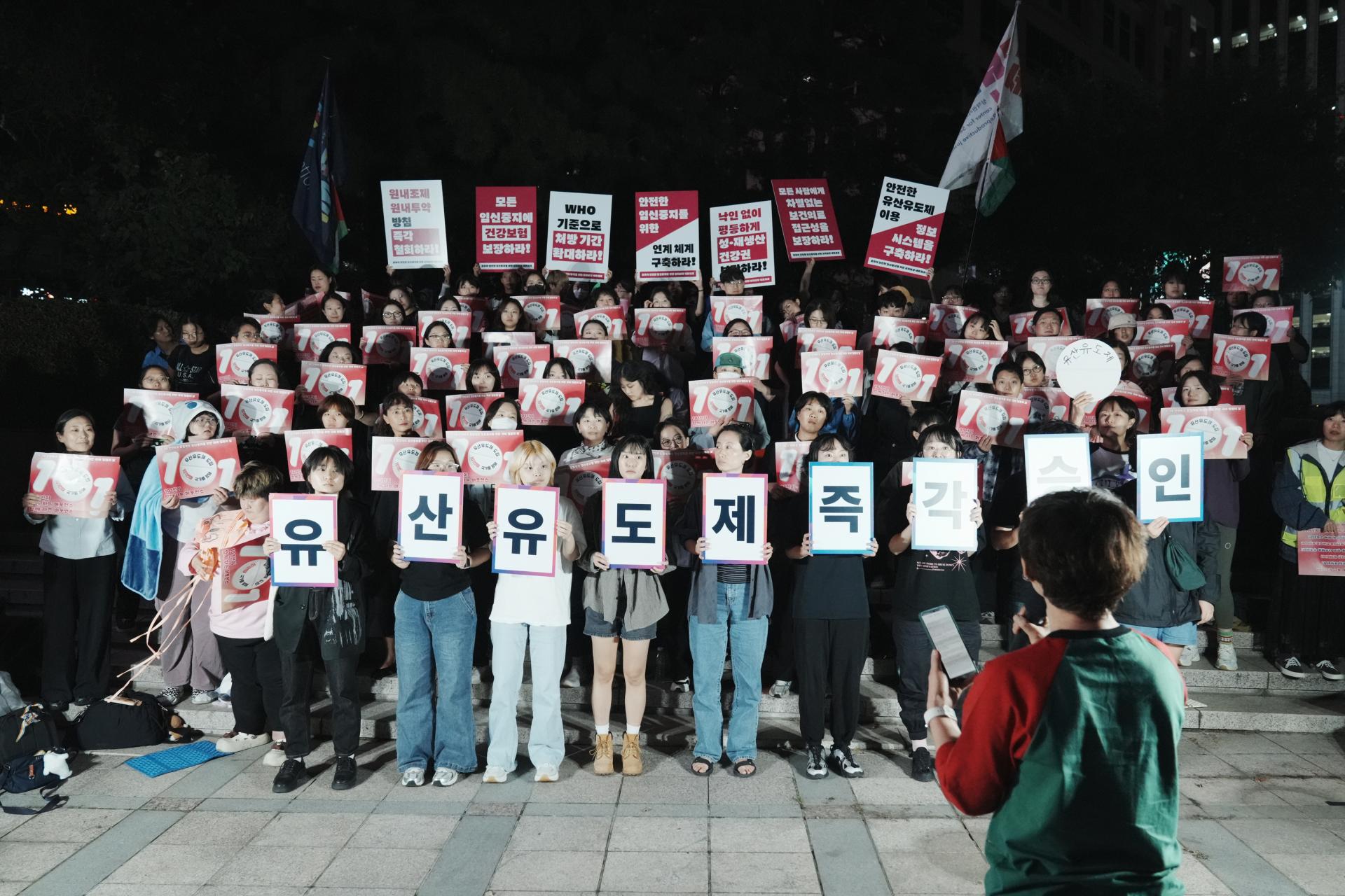 유산유도제라고 적힌 알약 모양의 피켓을 든 민우회 활동가가 사회자를 바라보고 있다. 다른 참여자들은 각자 피켓을 들고 유산유도제 알약 모형을 바라보며 그것이 건네질 순간을 기다리는 중이다. 사진 앞쪽으로는 몽실(사회자)의 뒷모습이 보인다. 유산유도제가 다음 순서를 향해 나아가기 직전의 모습.