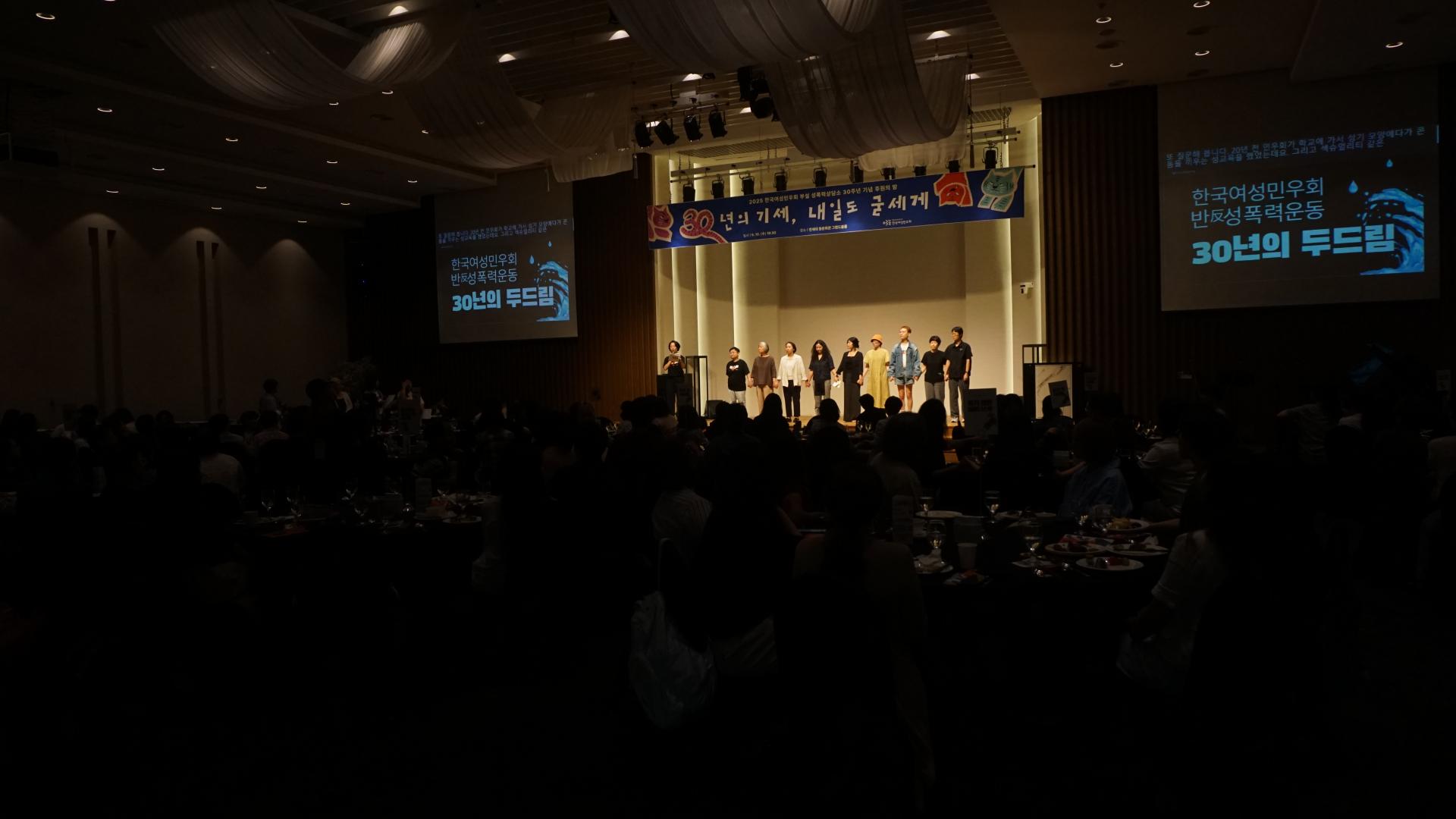 무대에 나란히 선 사람들과 사회자의 모습이 좀 더 멀리 찍힌 사진이다. 어둠 속 관객석의 많은 사람들의 실루엣이 이미지의 대부분을 차지한다. 무대 위가 노란 조명으로 밝게 빛나고 있고, 양 옆의 스크린에는 '한국여성민우회 반성폭력 운동 30년의 두드림'이라는 제목이 적혀 있다. 
