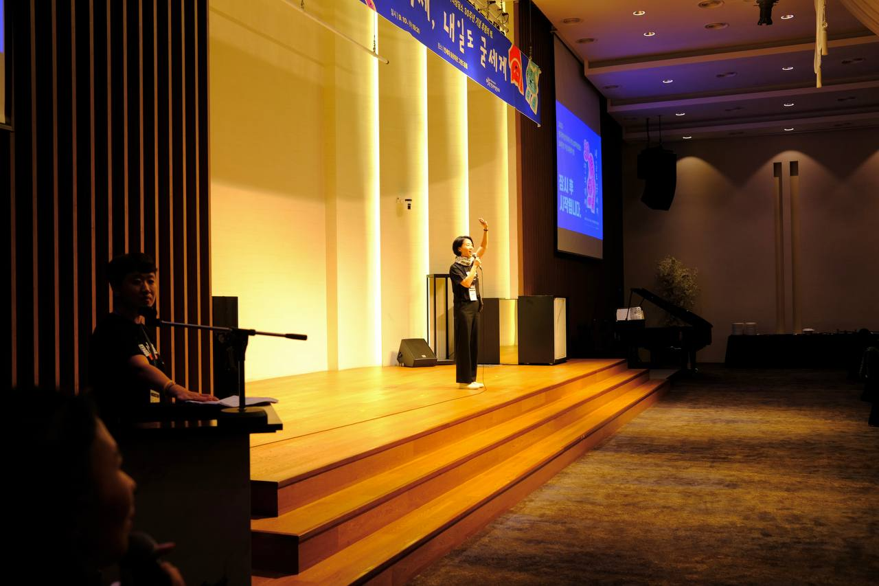 "밝게 조명된 무대 위에서 한 사람이 마이크를 들고 관객을 향해 손을 들어 인사하고 있다. 검은 셔츠와 검은 바지를 입고 흰 운동화를 신었다. 무대 위 배경에는 ‘30년의 기세, 내일도 굳세게’라는 파란색 현수막이 걸려 있고, 무대 오른쪽 벽에는 파란색 화면이 투사되어 안내 문구가 보인다. 객석은 어둡게 조명되어 있다."