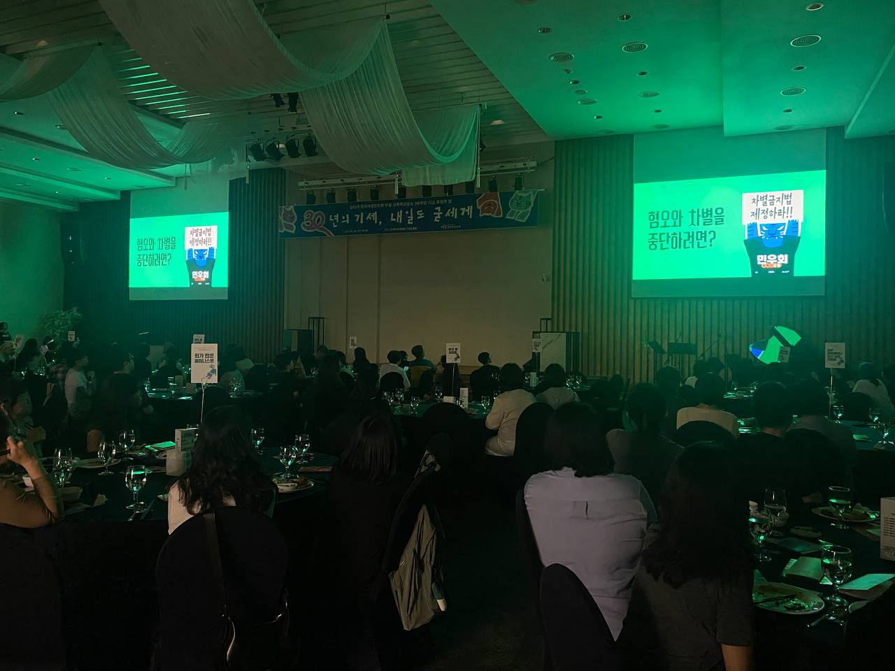 어둠 속에서 영상을 감상하는 사람들의 모습. 스크린에는 초록색 배경에 '혐오와 차별을 중단하려면?' '차별금지법 제정하라'라는 글자가 보인다. 