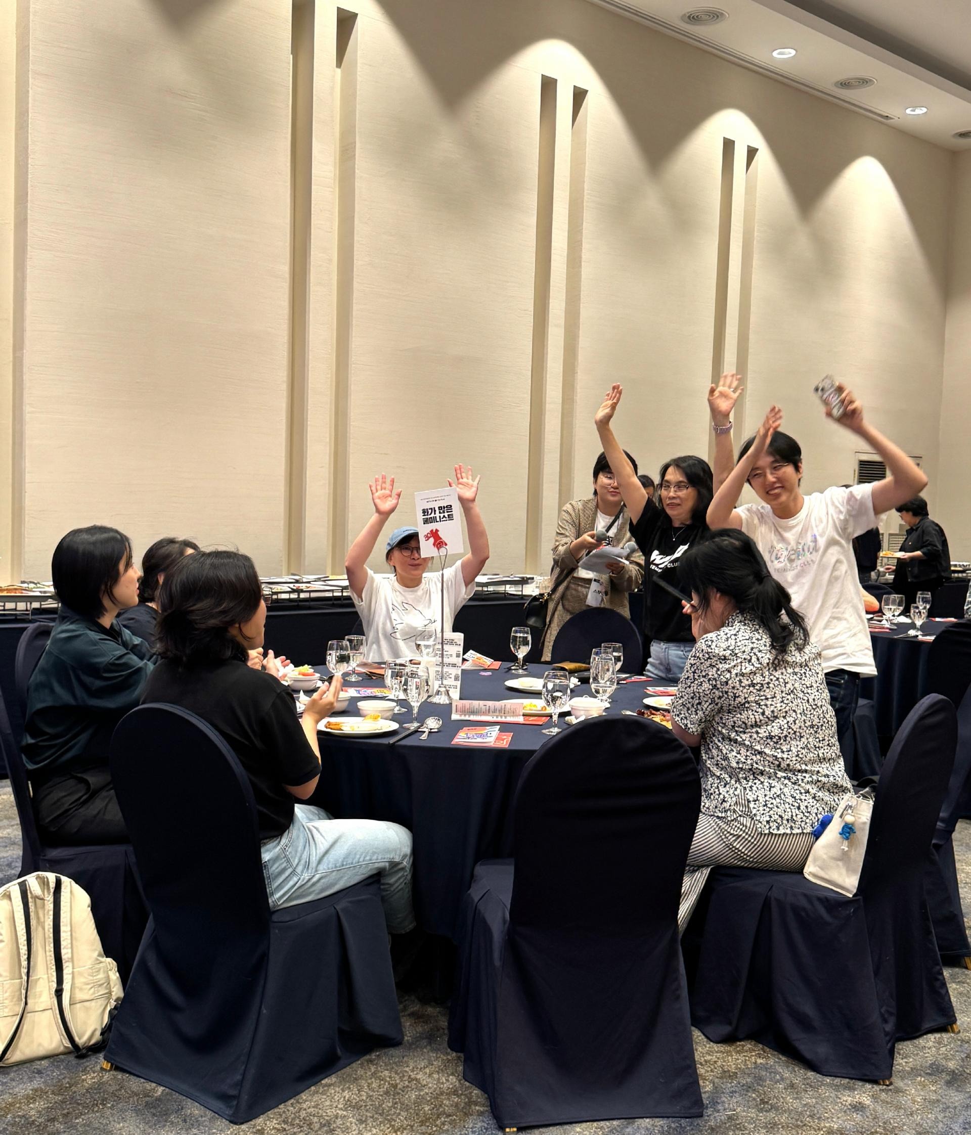 한 원탁의 구성원들이 청중을 향해 인사하는 모습. 세 명이 양손을 들고, 그 중 두 명은 자리에서 일어나 환히 웃으며 인사하는 모습이다. 