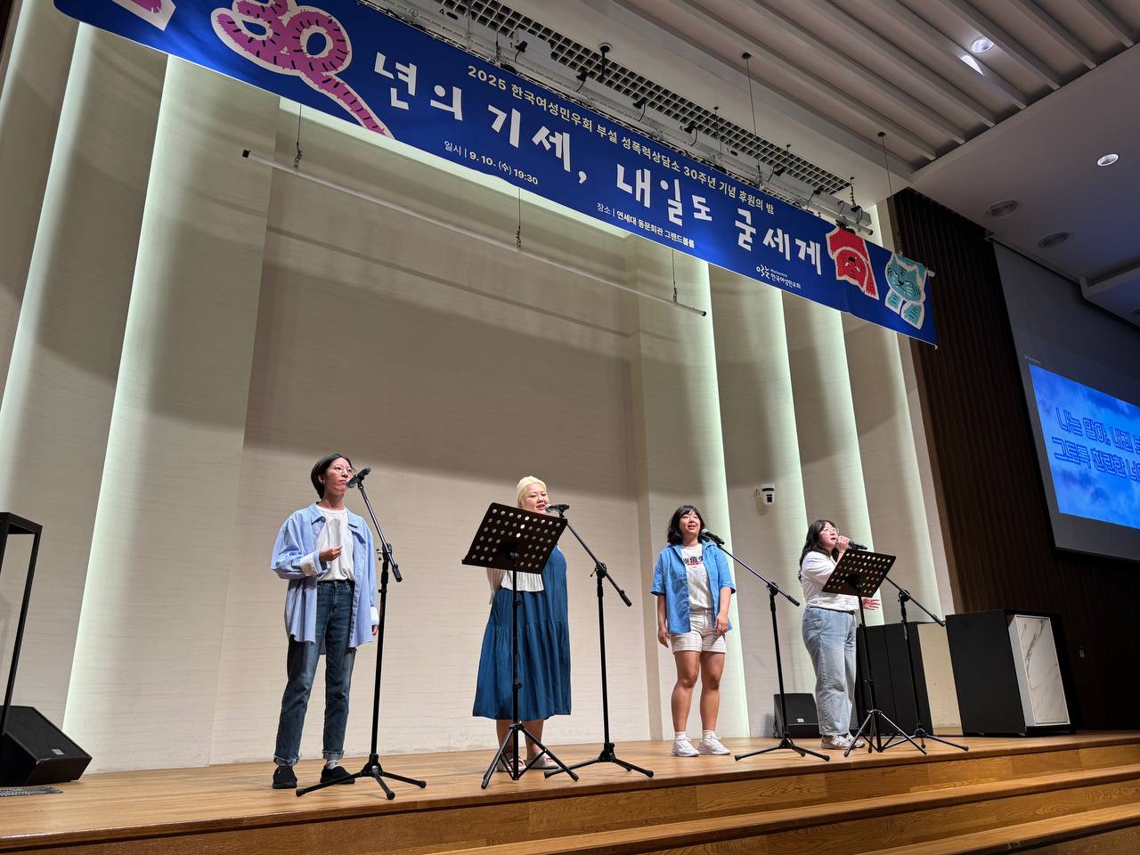 "밝게 조명된 무대 위에서 네 명의 여성이 마이크 앞에 서 있다. 모두 파란색이나 흰색 계열의 옷을 입었고, 노래하거나 발언하는 듯 표정이 밝다. 무대 위에는 ‘30년의 기세, 내일도 굳세게’라고 적힌 파란 현수막이 걸려 있고, 오른쪽 벽에는 파란색 배경의 가사 화면이 빔프로젝터로 비춰지고 있다."