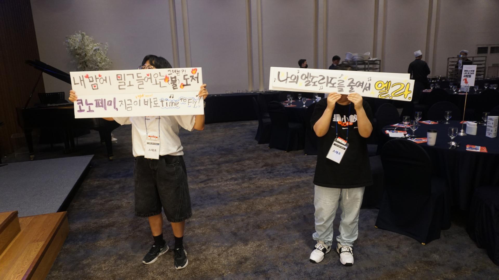 무대 위 공연팀을 향해 피켓을 흔들고 있는 두 사람의 모습. "내 맘에 밀고 들어오는 그대는 불도저", "코노페미, 지금이 바로 time to fly", "나의 엘도라도를 줄여서 엘라"라고 적혀 있다. 