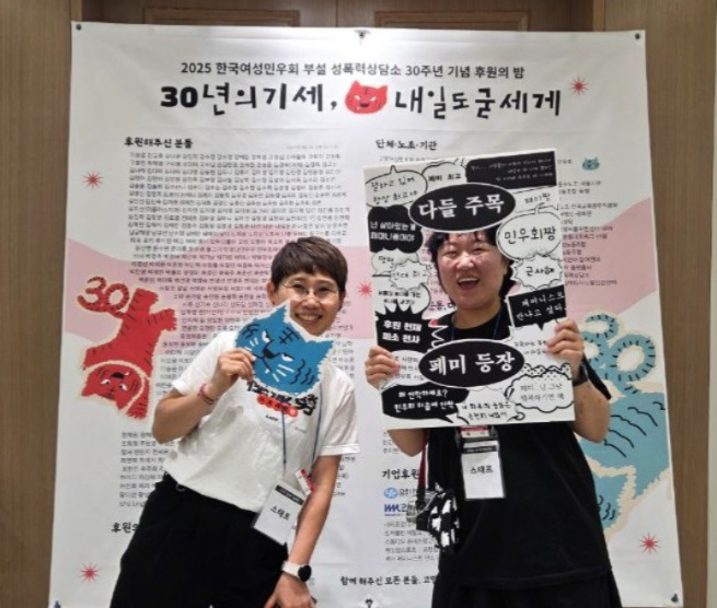 후원자 명단이 적힌 행사 현수막을 배경으로 두 명의 활동가가 환하게 웃고 있는 모습. 한 명은 얼굴을 내밀 수 있는 피켓을, 한 명은 파란색 호랑이얼굴 모양의 피켓을 들고 있다. 