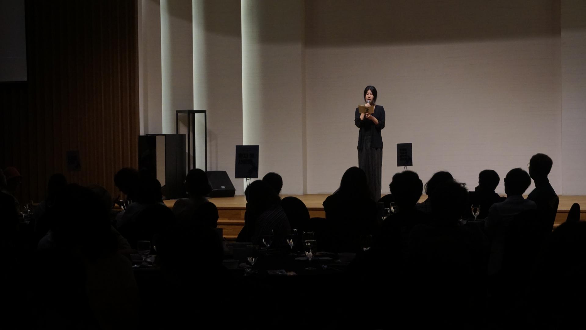 "무대 한가운데에서 검은 옷을 입은 사람이 마이크를 들고 원고를 읽으며 발언하고 있다. 무대는 밝게 조명되어 있고, 객석은 어둡게 처리되어 사람들의 실루엣만 보인다. 테이블 위에는 물컵과 프로그램 책자가 놓여 있다."