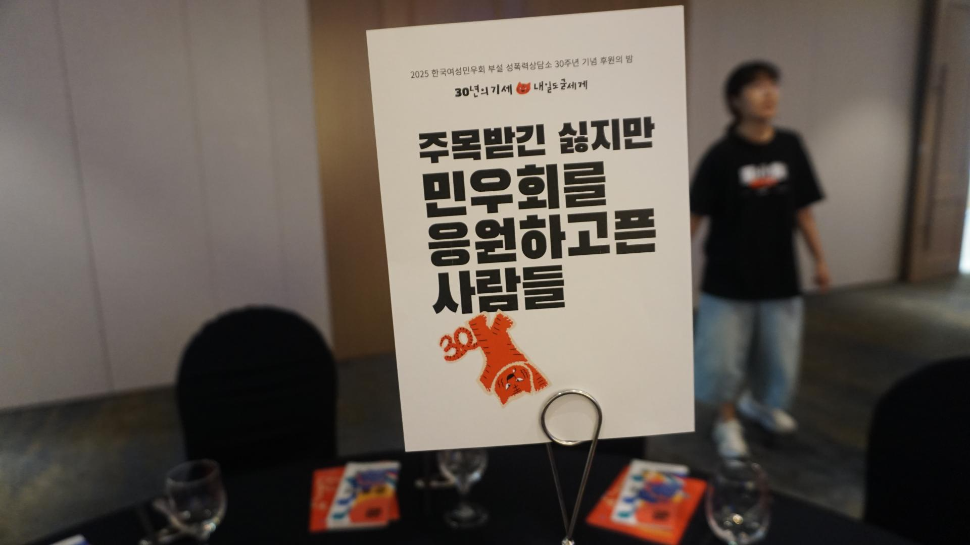 행사장 테이블 위 팻말: '주목받긴 싫지만 민우회를 응원하고픈 사람들'. 빨간색 호랑이가 거꾸로 선 이미지가 들어가 있다.