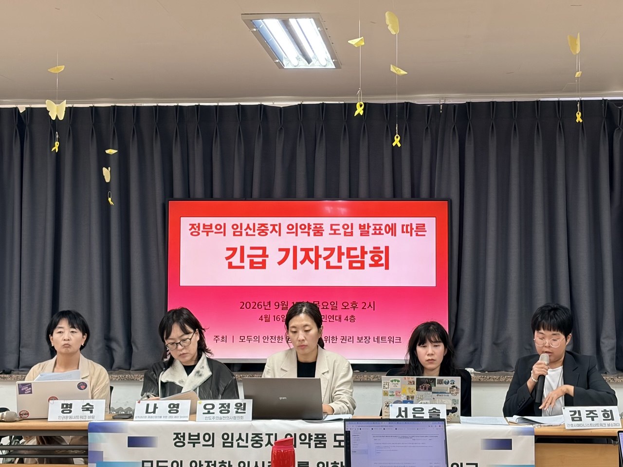 다섯명의 사람이 기자간담회 제목이 적힌 텔레비전 앞에 앉아 노트북 또는 인쇄물을 보고 있다. 우측 끝에 앉은 사람이 마이크를 들고 발언 중이다.