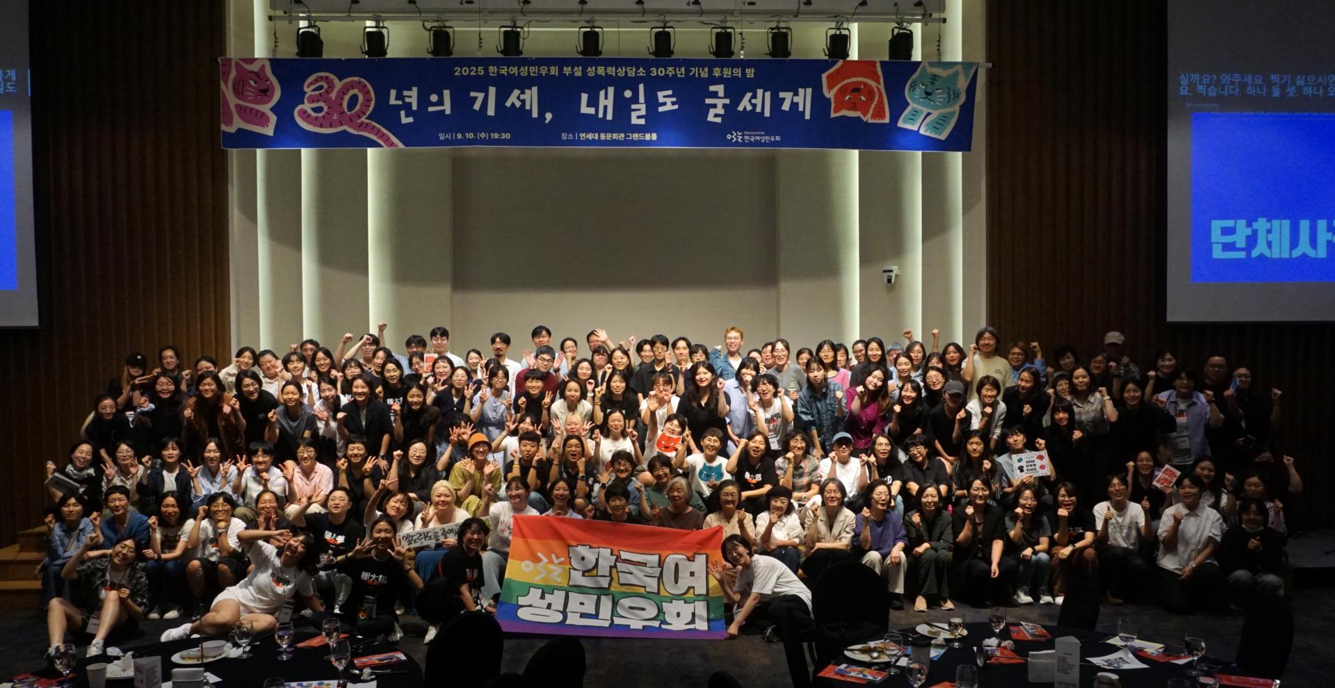 실내 행사장에서 150여 명이 무대 위에 모여 단체사진을 찍고 있다. 가운데에 무지개 색 배너를 든 사람들이 있고, 배너에는 '한국여성민우회'라는 글자가 적혀 있다. 무대 뒤에는 파란색 현수막이 걸려 있으며, ‘30년의 기세, 내일도 굳세게’라는 문구와 함께 빨간색과 파란색 그림이 보인다. 무대 앞쪽 테이블 위에는 식사 접시와 음료가 놓여 있다.
