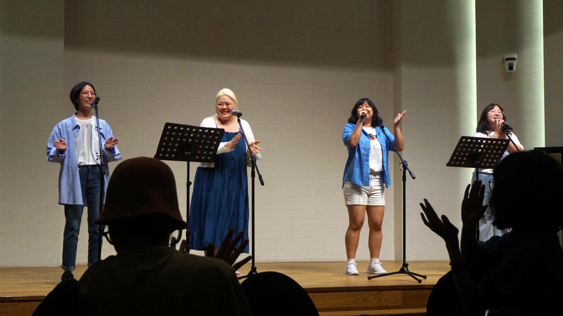 "무대 위에 네 명의 여성이 마이크 앞에 서서 노래하거나 말하고 있다. 모두 파란색이나 흰색 계열의 옷을 입었고, 두 사람은 손뼉을 치거나 손짓하며 즐겁게 공연 중이다. 앞쪽 객석에는 관객들이 손을 들고 호응하고 있다."