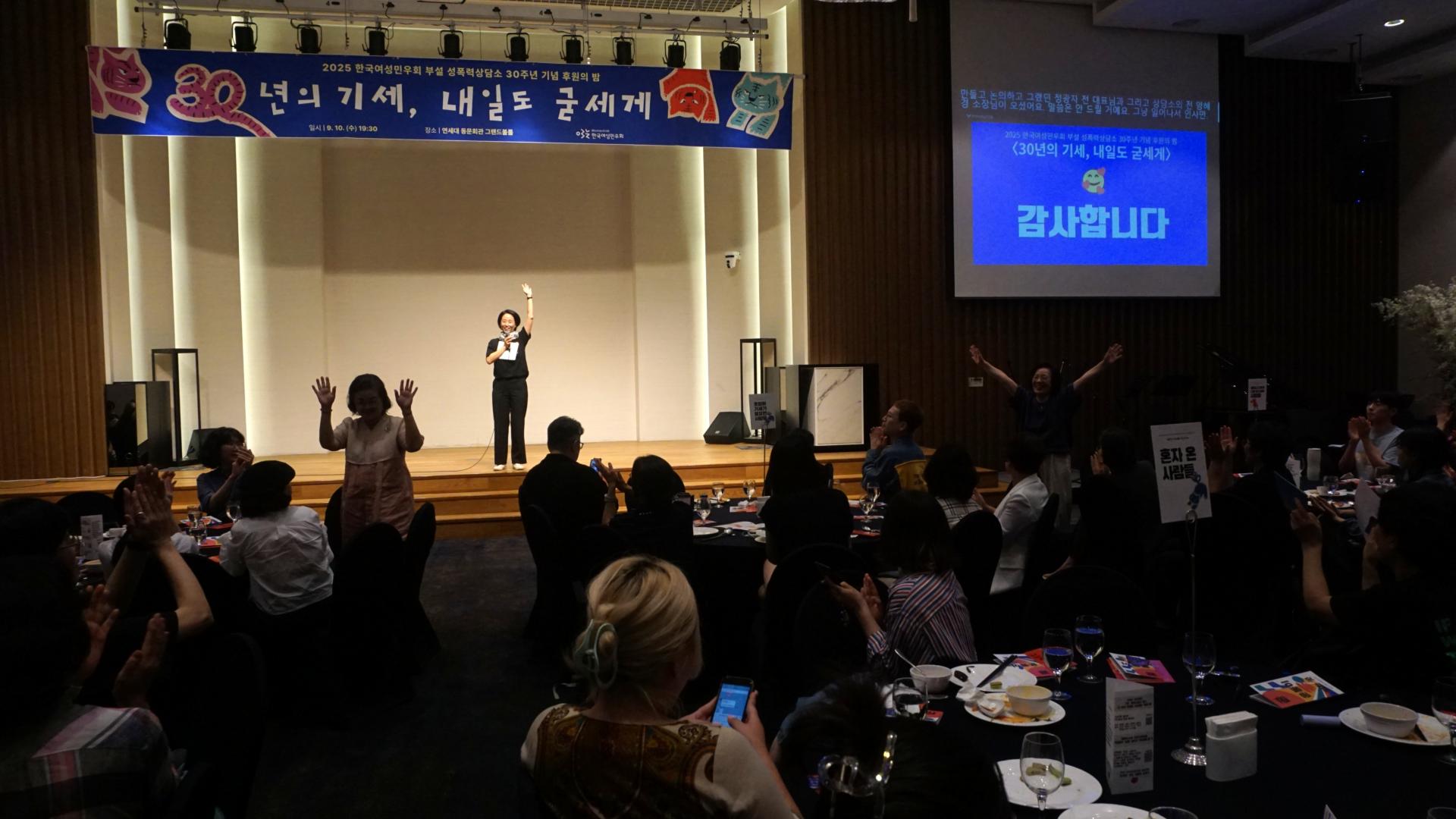 "밝은 무대 위에서 검은 옷을 입은 사람이 마이크를 들고 한 손을 높이 들며 인사하고 있다. 무대 위에는 ‘30년의 기세, 내일도 굳세게’라고 적힌 파란색 현수막이 걸려 있다. 객석에 앉아 있던 사람들이 박수를 치거나 손을 흔들고, 두 명이 일어나서 두 팔을 들고 환호하고 있다. 오른쪽 스크린에는 파란색 배경에 ‘감사합니다’라는 글자가 크게 비춰지고 있다."