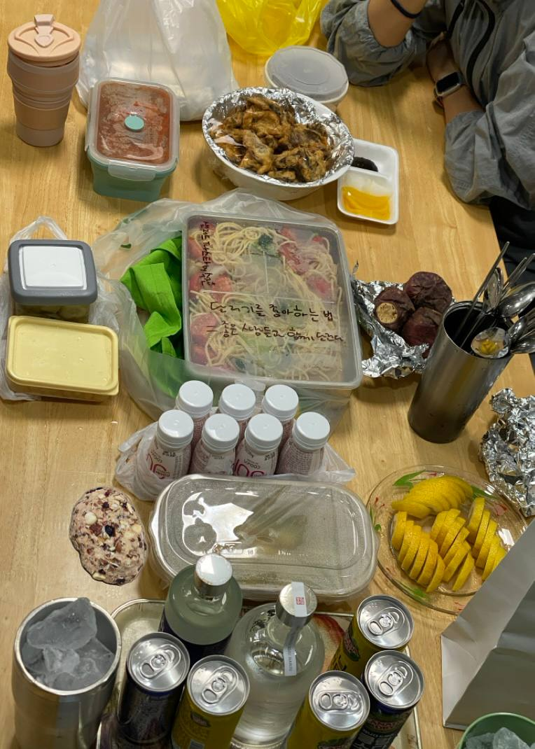 책상 위에 버섯 튀김, 샐러드 파스타, 떡볶이 등 먹을 거리가 늘여져있다. 그 옆엔 얼음이 담긴 컵과 술, 탄산음료, 레몬 등이 있다.