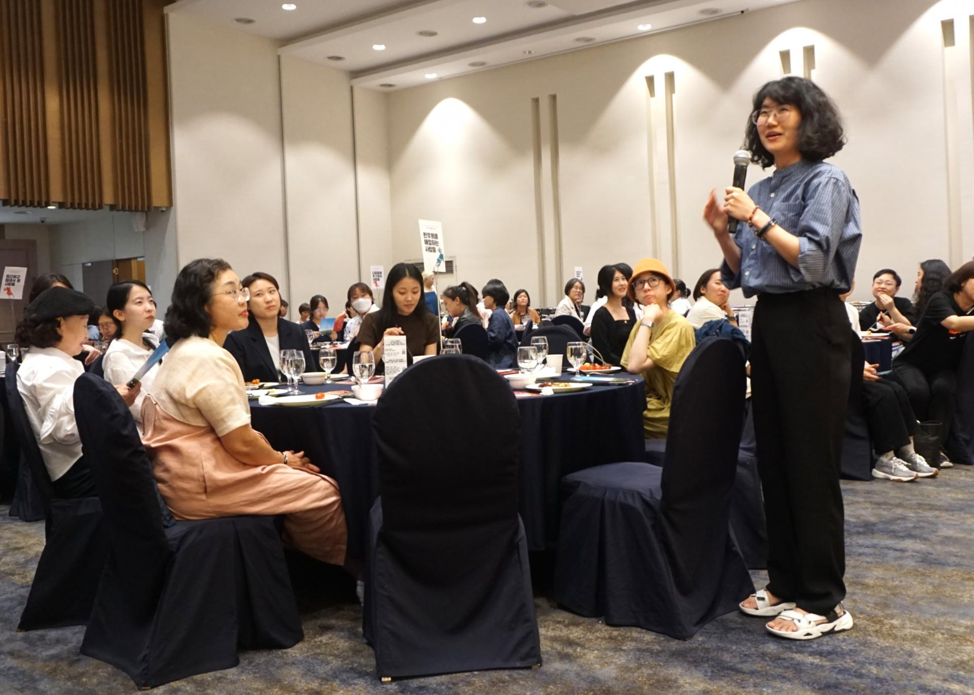 여러 청중들이 원탁에 앉아 바라보는 가운데, 푸른 셔츠를 입고 단발머리를 한 사람이 일어서서 마이크를 들고 웃으며 말하고 있는 모습. 