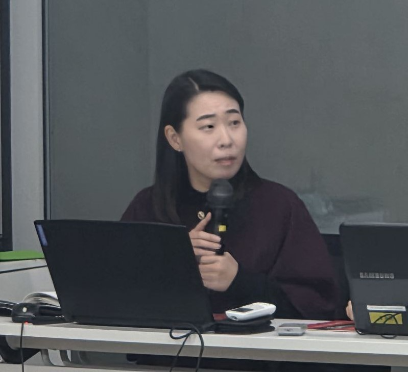 노트북 앞에 긴 머리의 여성이 마이크를 들고 발표를 진행하고 있는 모습이다.