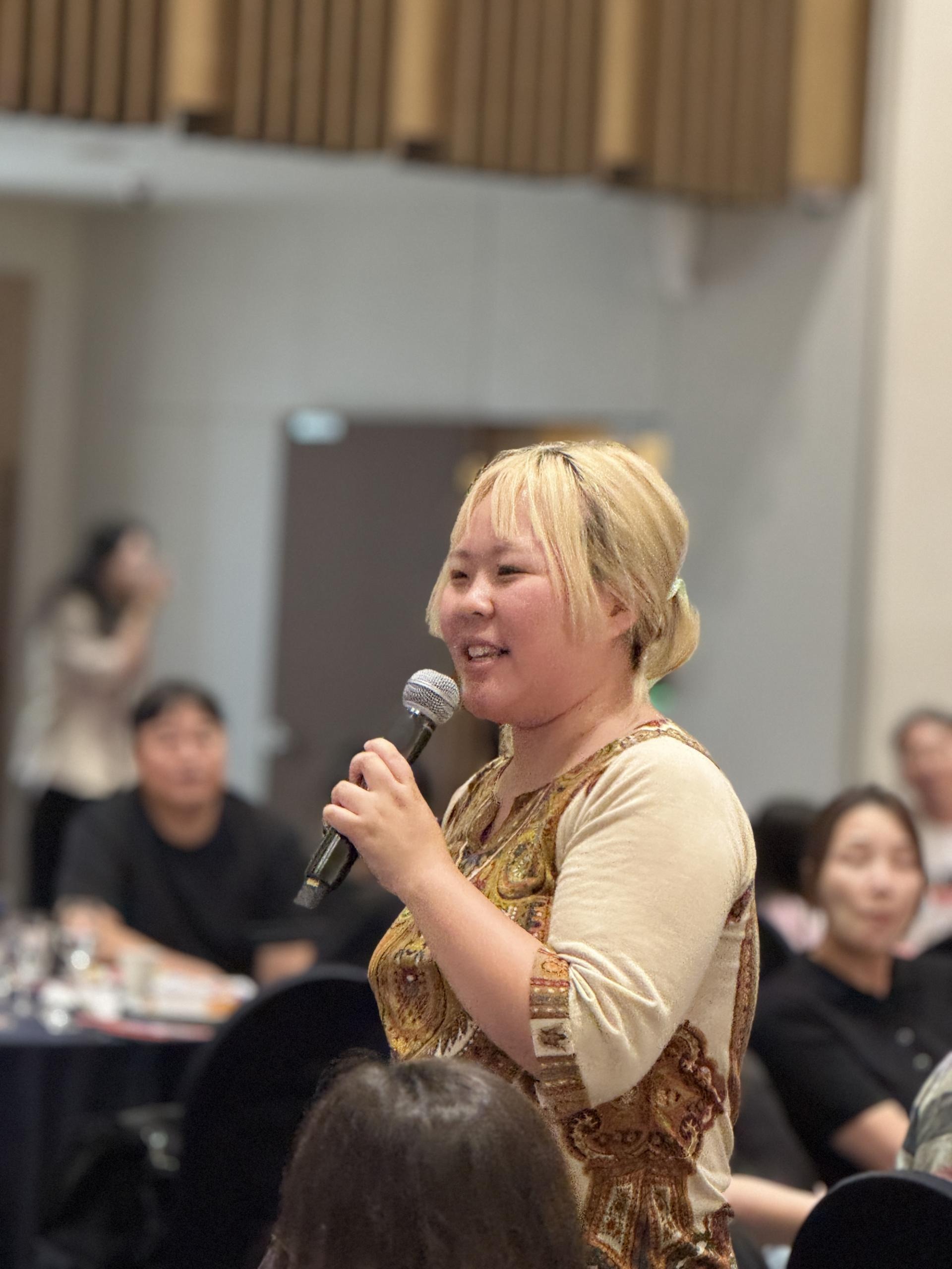 청중 가운데에 선, 노란색으로 탈색된 머리의 한 사람이 마이크를 들고 웃으며 말하고 있는 모습 