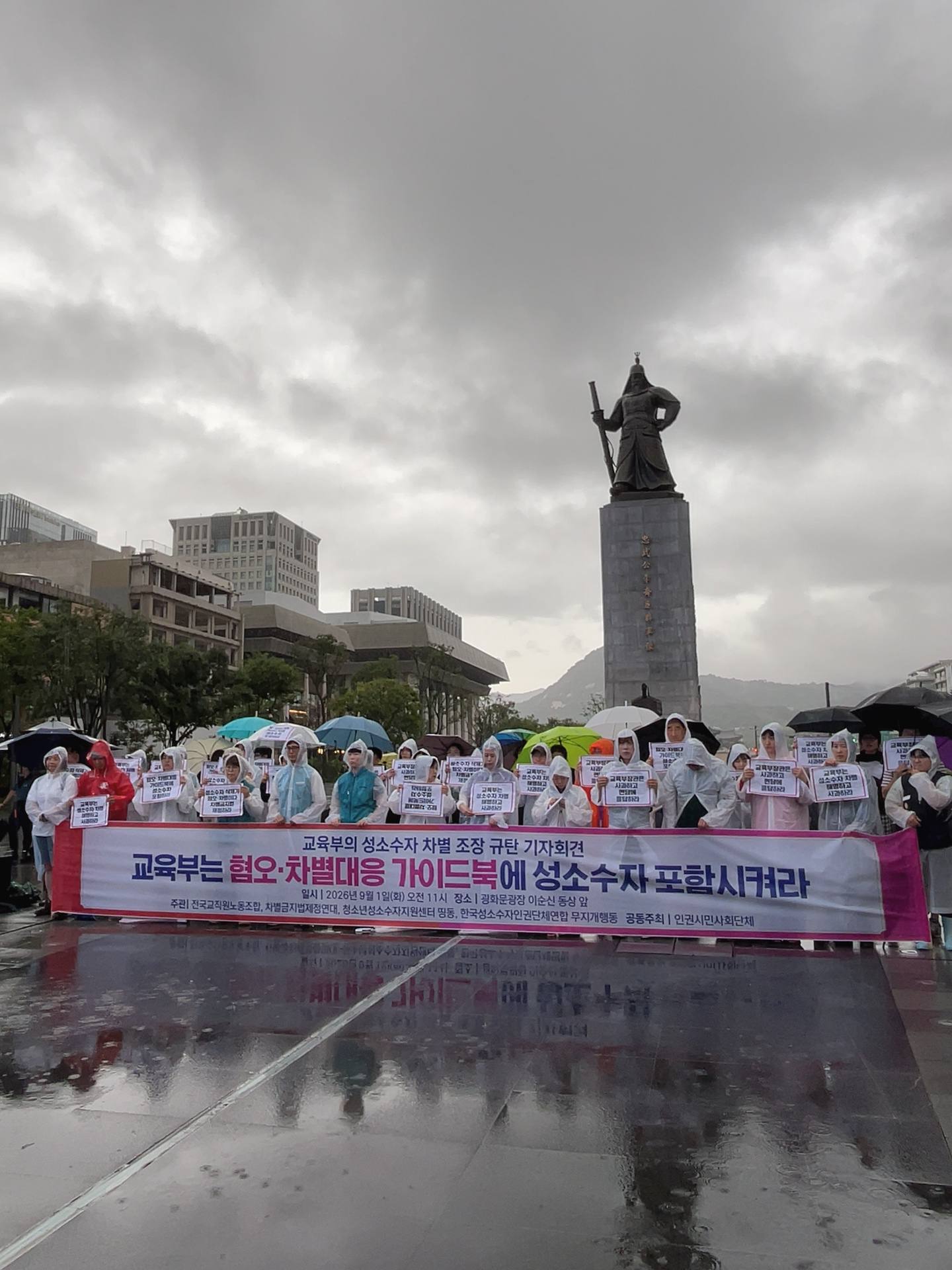 광화문 광장 이순신 동상 앞 쪽으로 사람들이 우비를 입거나 우산을 든 채 서 있고, 교육부를 규탄하며 가이드북에 성소수자 포함시키라는 요구가 담긴 피켓과 현수막을 들고 있다