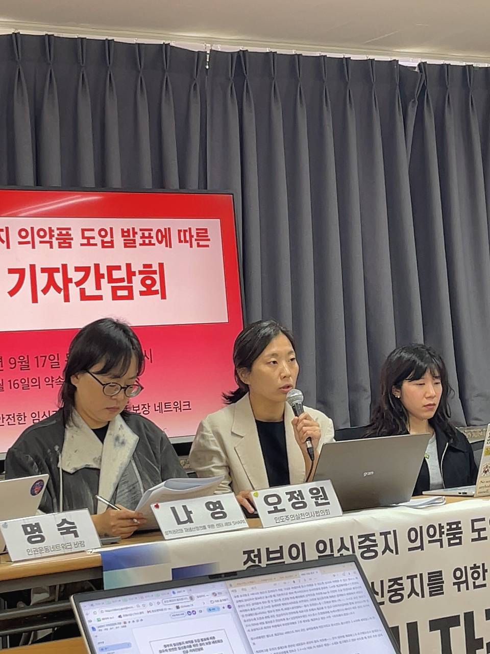 한 사람이 마이크를 들고 발언 중이다. 그 옆으로는 네 명의 사람이 앉아서 자료를 보고 있고, 뒤로는 기자간담회 제목이 적힌 텔레비전이, 책상 앞쪽에는 기자간담회 제목이 적힌 현수막이 걸려 있다.