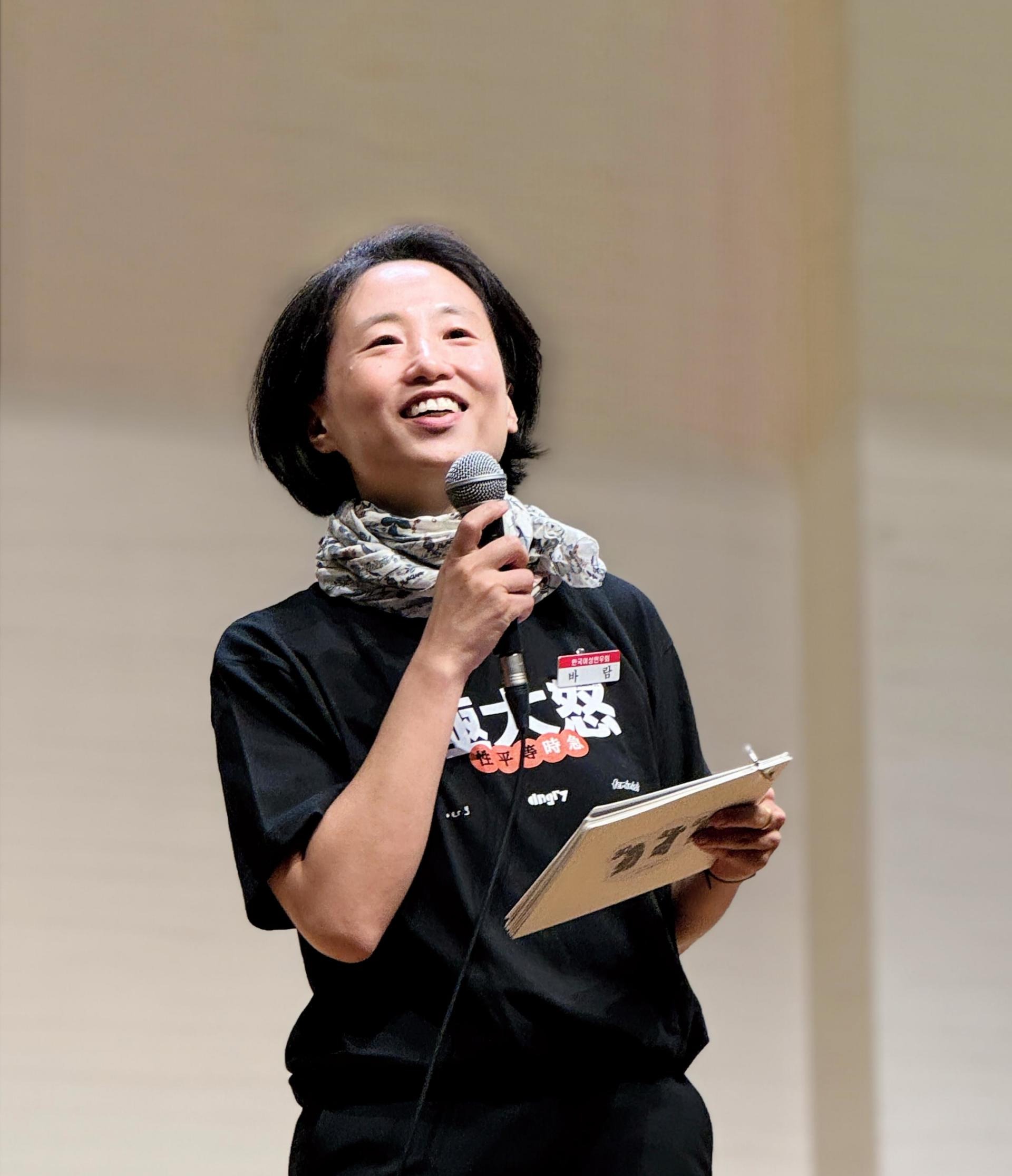 사회자가 마이크와 대본이 적힌 노트를 들고 환하게 웃고 있는 모습. 