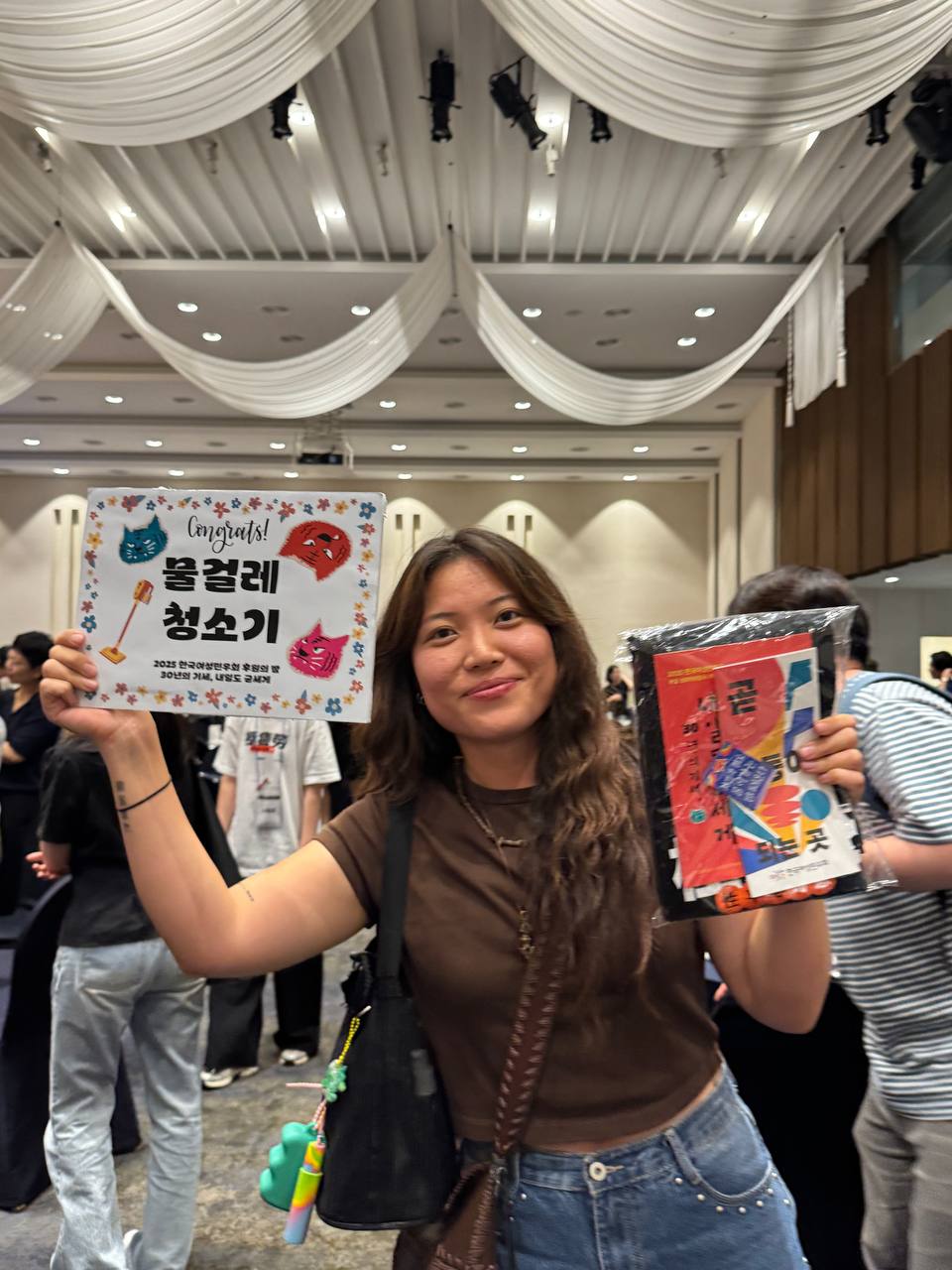 갈색 반팔 티셔츠를 입고 긴 머리를 한 사람이 '물걸레 청소기'라고 적힌 A4용지 크기의 종이를 들어보이며 미소짓고 있는 모습. 