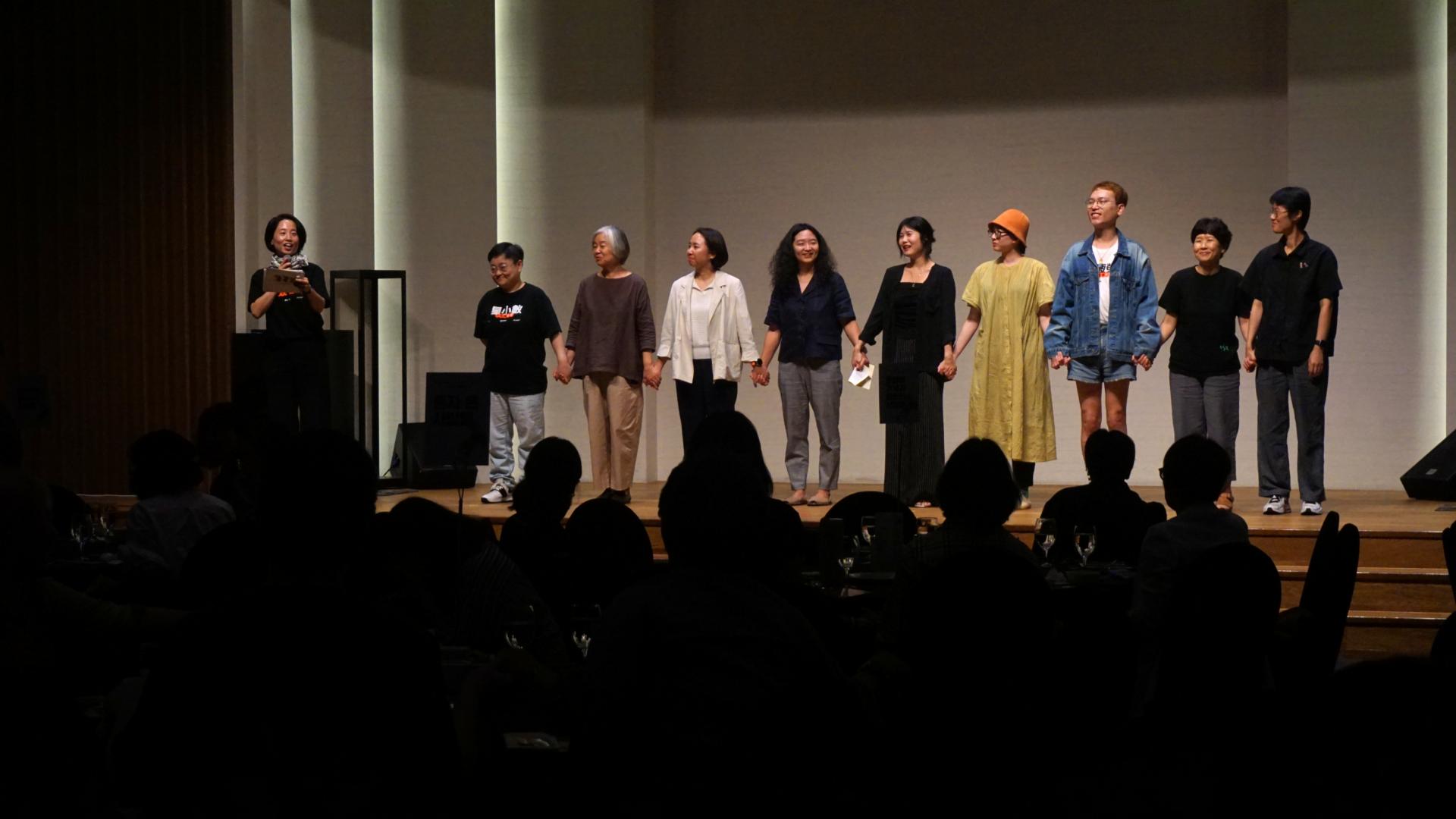 "무대 위에 10명이 나란히 서서 서로 손을 잡고 관객을 향해 서 있다. 왼쪽 끝에는 마이크를 들고 진행 중인 사람이 있고, 나머지 사람들은 다양한 옷차림으로 서 있으며 웃고 있다. 객석은 어둡게 비쳐 사람들의 실루엣만 보인다."