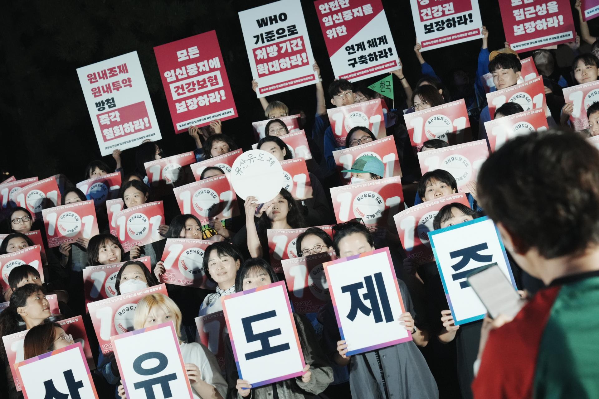 참여자들의 손과 손을 통해 유산유도제 알약 모형이 건네지고 있는 모습. 유산유도제 즉각승인이라는 작은 직사각형 모양의 피켓을 든 활동가 뒤쪽으로 선 참여자들은 손피켓을 들고 있다. 모두 알약 모형을 이어받기 위해 순서를 기다리는 중이다. 사진 앞쪽에는 몽실의 뒷모습이 보이고 있으며, 몽실은 휴대폰을 보며 사회를 이어가고 있다.