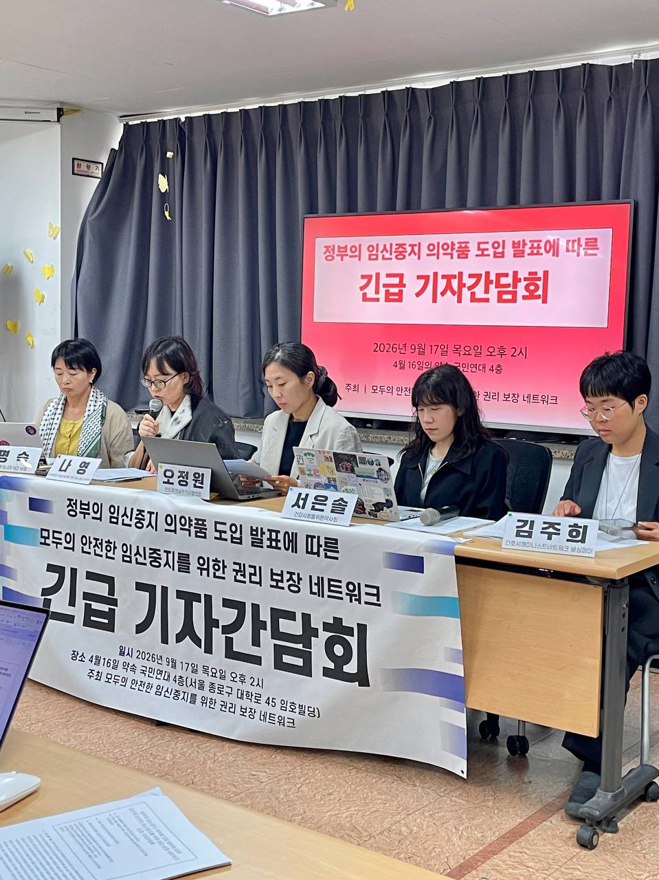 한 사람이 마이크를 들고 발언 중이다. 그 옆으로는 네 명의 사람이 앉아서 자료를 보고 있고, 뒤로는 기자간담회 제목이 적힌 텔레비전이, 책상 앞쪽에는 기자간담회 제목이 적힌 현수막이 걸려 있다.