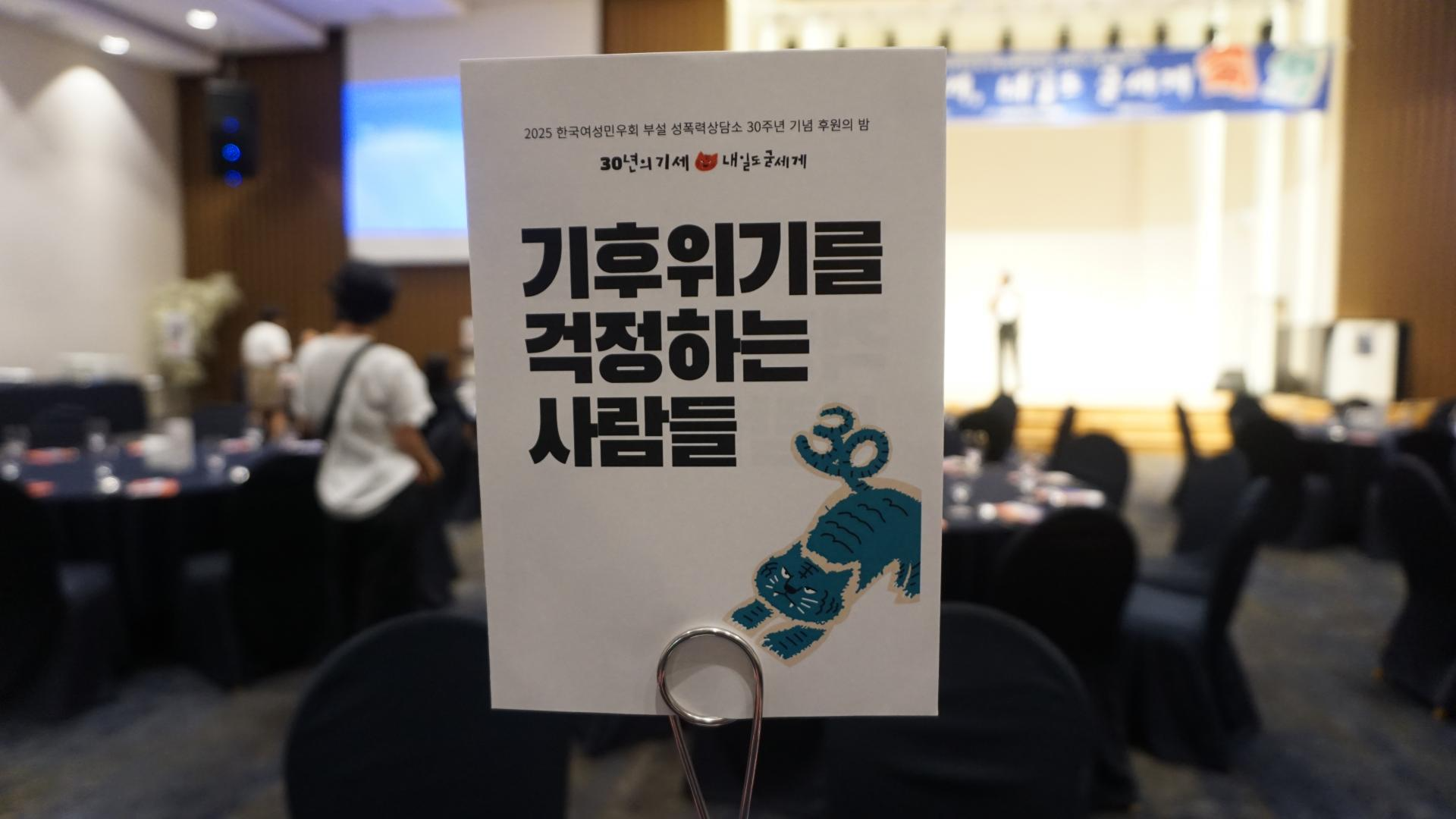 행사장 테이블 위 팻말: '기후위기를 걱정하는 사람들'. 파란색 호랑이가 팔을 앞으로 쭉 빼고 누운 이미지가 들어가 있다.