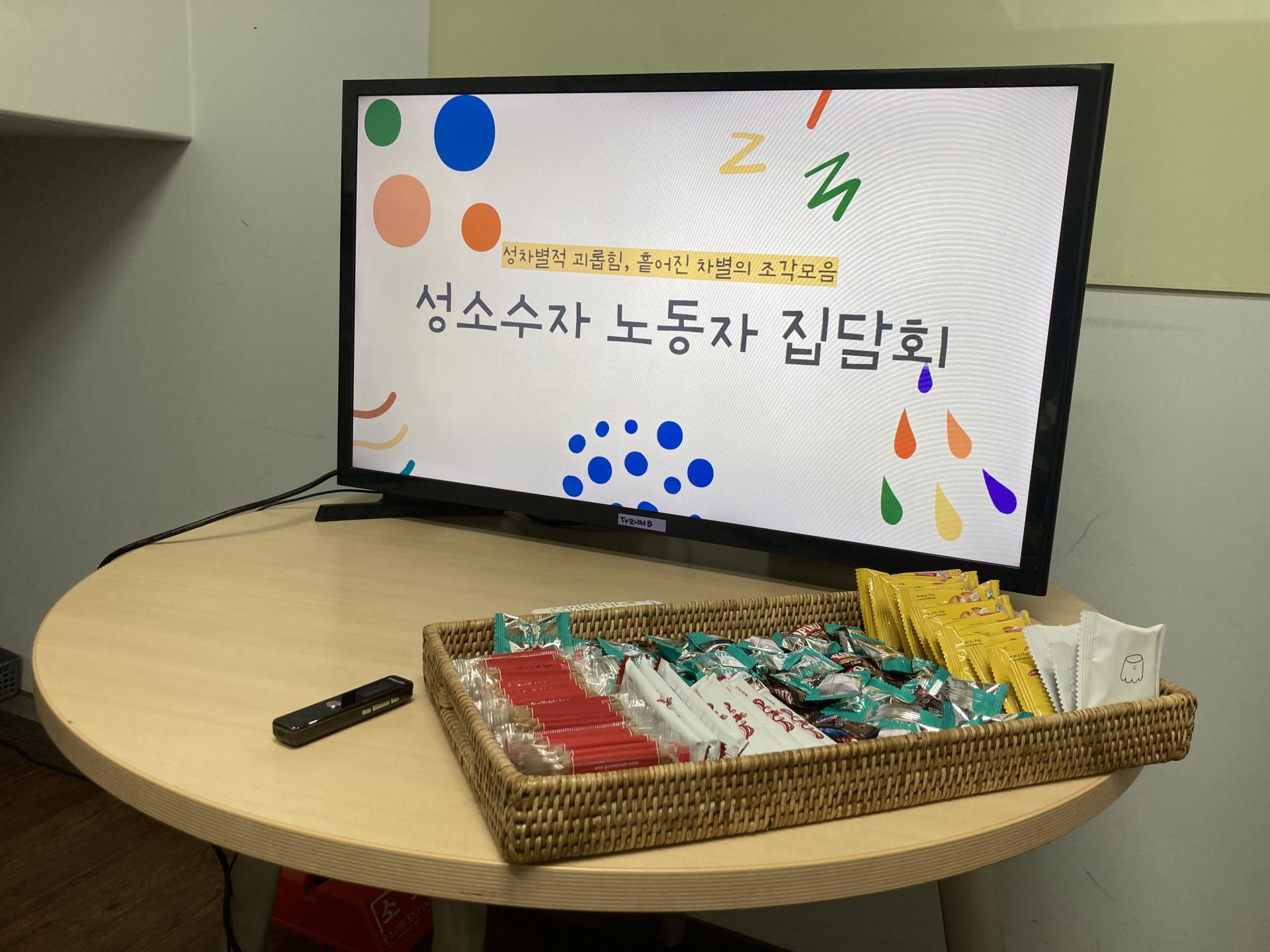 동그란 테이블 위에 큰 모니터가 있다. 모니터에는 "성차별적 괴롭힘, 흩어진 차별의 조각모음" "성소수자 노동자 집담회"라고 쓰여있다. 모니터 앞에는 다양한 과자가 갈색 바구니 안에 준비되어 있는 모습이다.