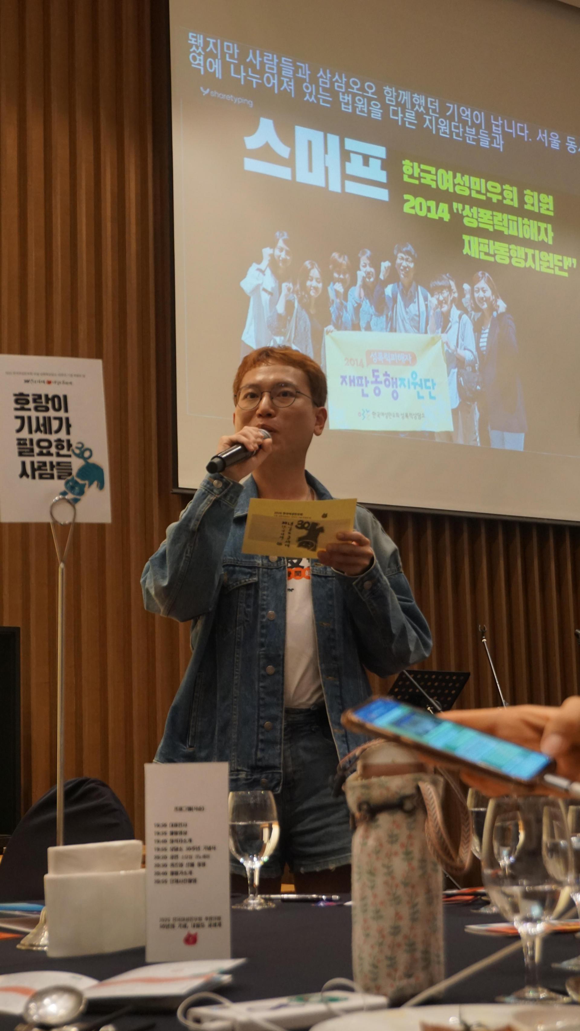 "짧은 머리를 한 사람이 청색 청재킷과 청반바지를 입고 무대 앞에서 마이크를 들고 발언하고 있다. 한 손에는 노란색 종이를 들고 있으며, 뒤쪽 스크린에는 ‘스머프 한국여성민우회 회원 2014 성폭력피해자 재판동행지원단’이라는 문구와 함께 사람들 단체사진이 크게 비춰지고 있다. 왼쪽에는 ‘호랑이 기세가 필요한 사람들’이라고 적힌 테이블 표지판이 세워져 있다. 테이블 위에는 와인잔, 휴대폰, 보온병, 메뉴 카드 등이 놓여 있다.