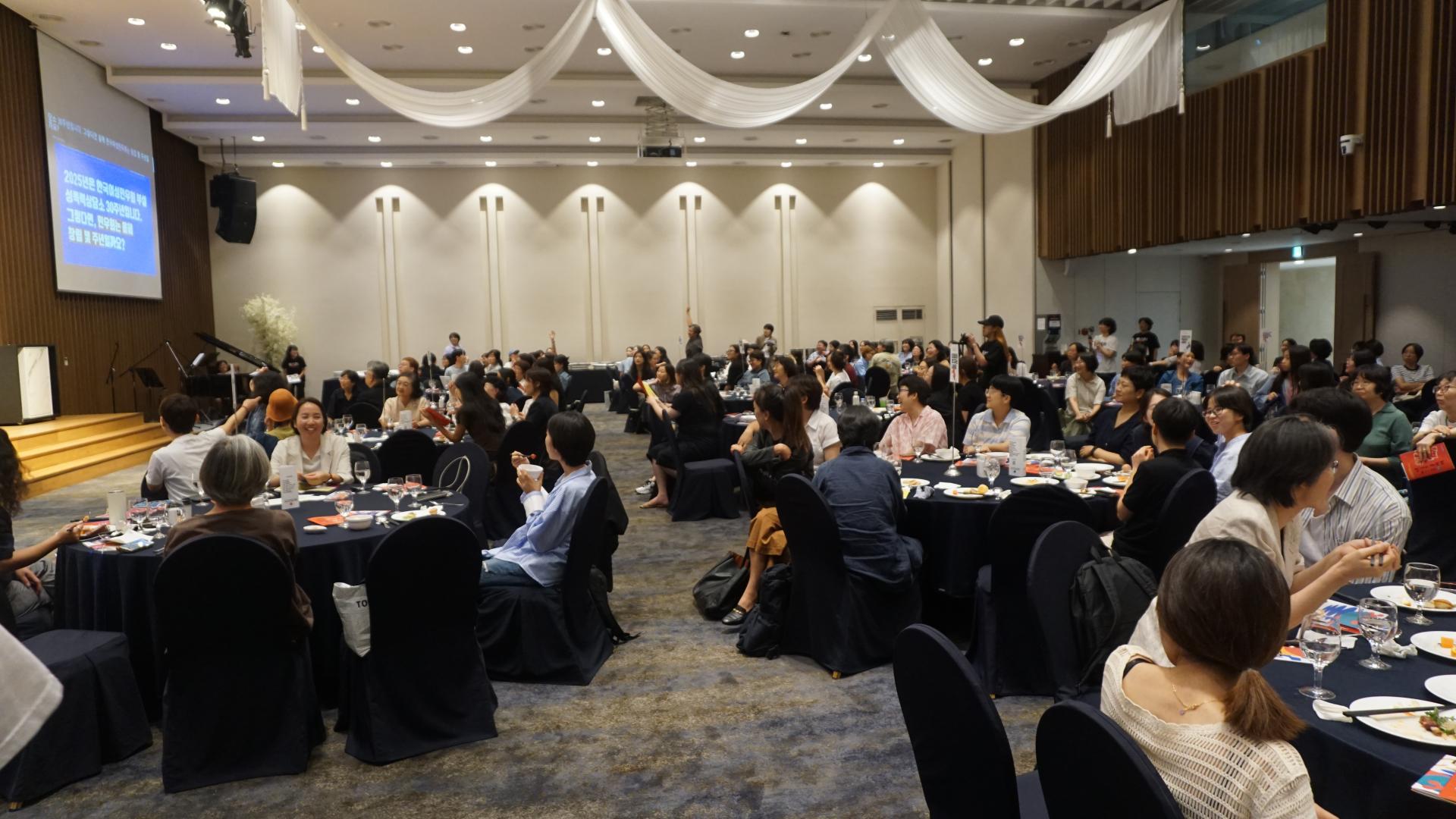행사장의 모습. 원형테이블들에 많은 사람이 앉아 있다. 웃고 있는 사람들, 군데군데 사람들이 한 손을 번쩍 들고 있는 모습이 보인다. 