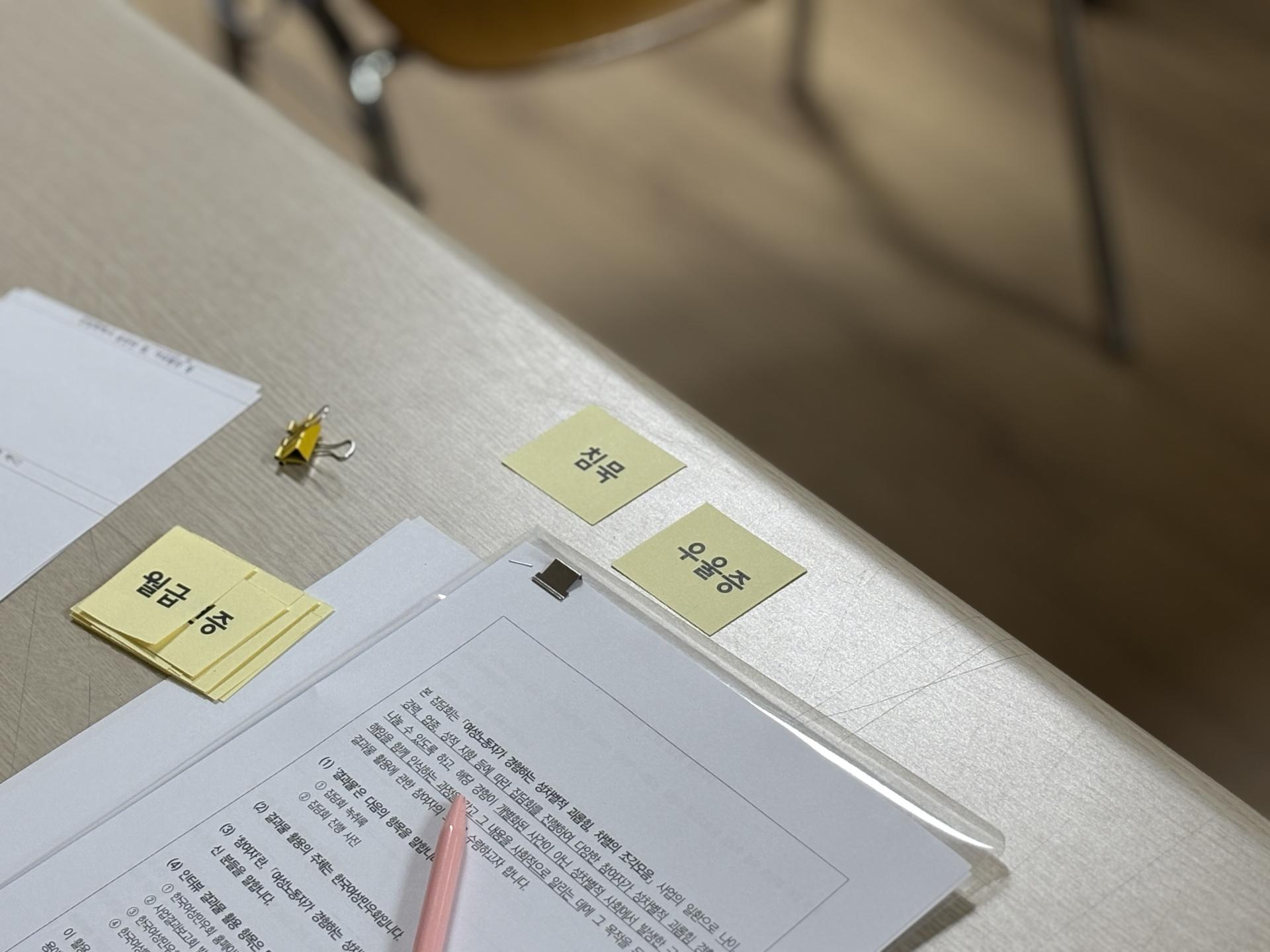 베이지색 테이블 위에 서류와 연노랑색 작은 종이조각들이 있다. 종이조각들에는 "월급", "침묵", "우울증" 등 성차별적 괴롭힘으로 인한 영향을 키워드로 한 문구들이 쓰여있다.