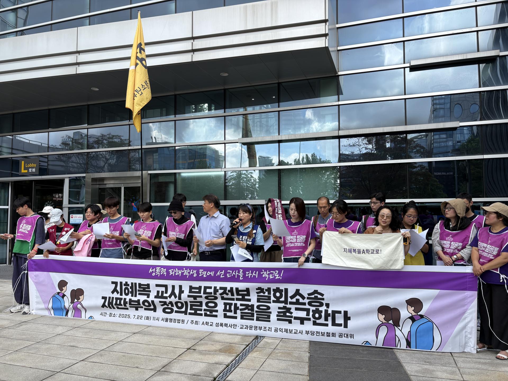 지혜복 교사 부당전보 철회소송을 촉구하기 위한 기자회견이 진행되고 있는 모습이다. 참여자들은 보라색 연대의 조끼를 입고 결연한 표정으로 서있다.