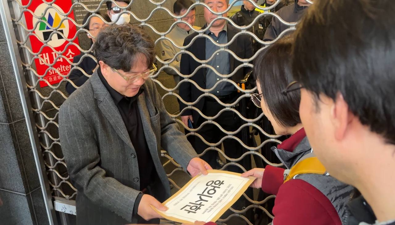 회색 재킷을 입은 남자가 항의서한이라 쓰여진 서류봉투를 받고 있다.