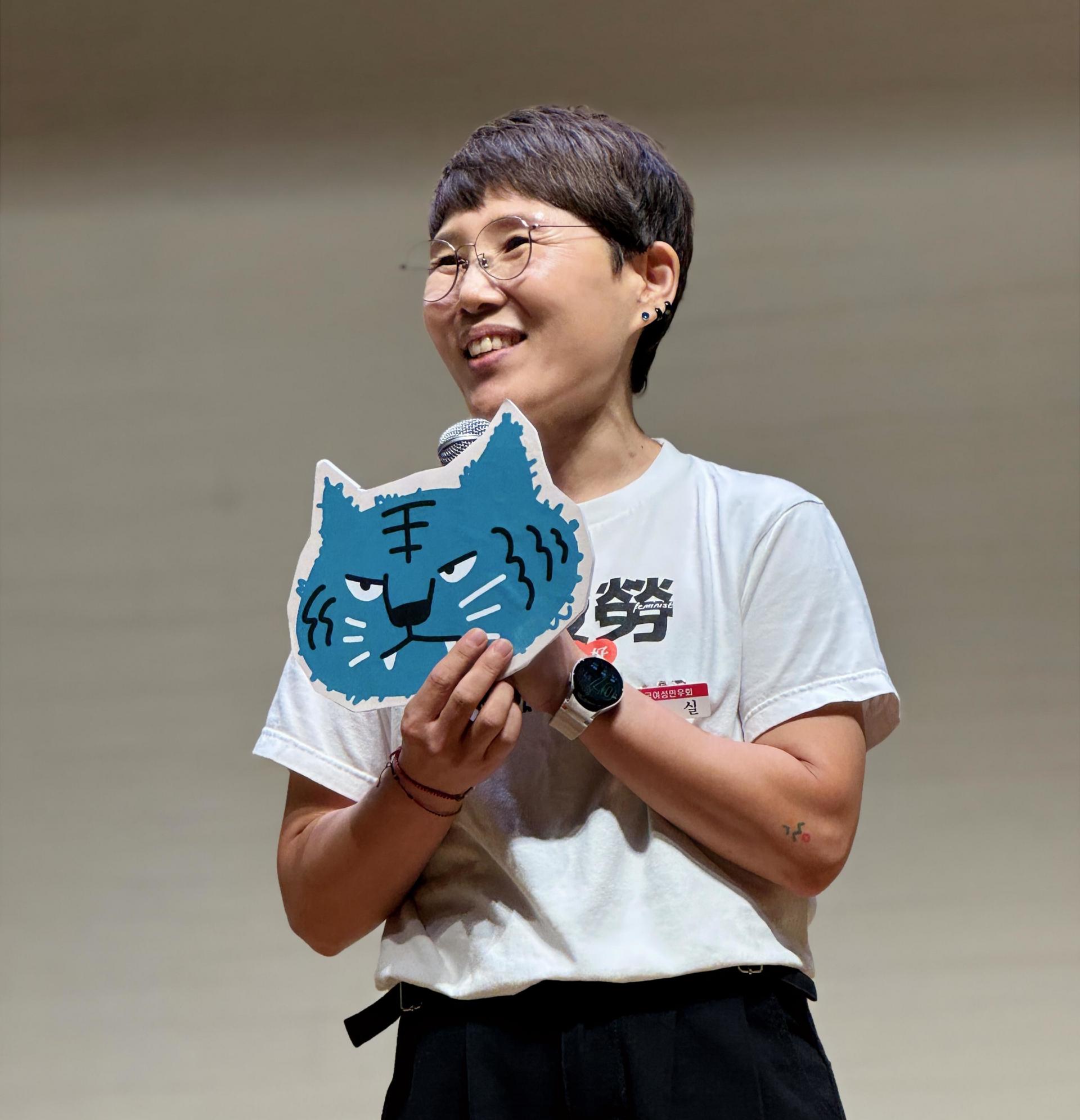 파란색 호랑이 피켓을 든 사람이 마이크를 들고 웃으며 뭔가를 말하는 모습