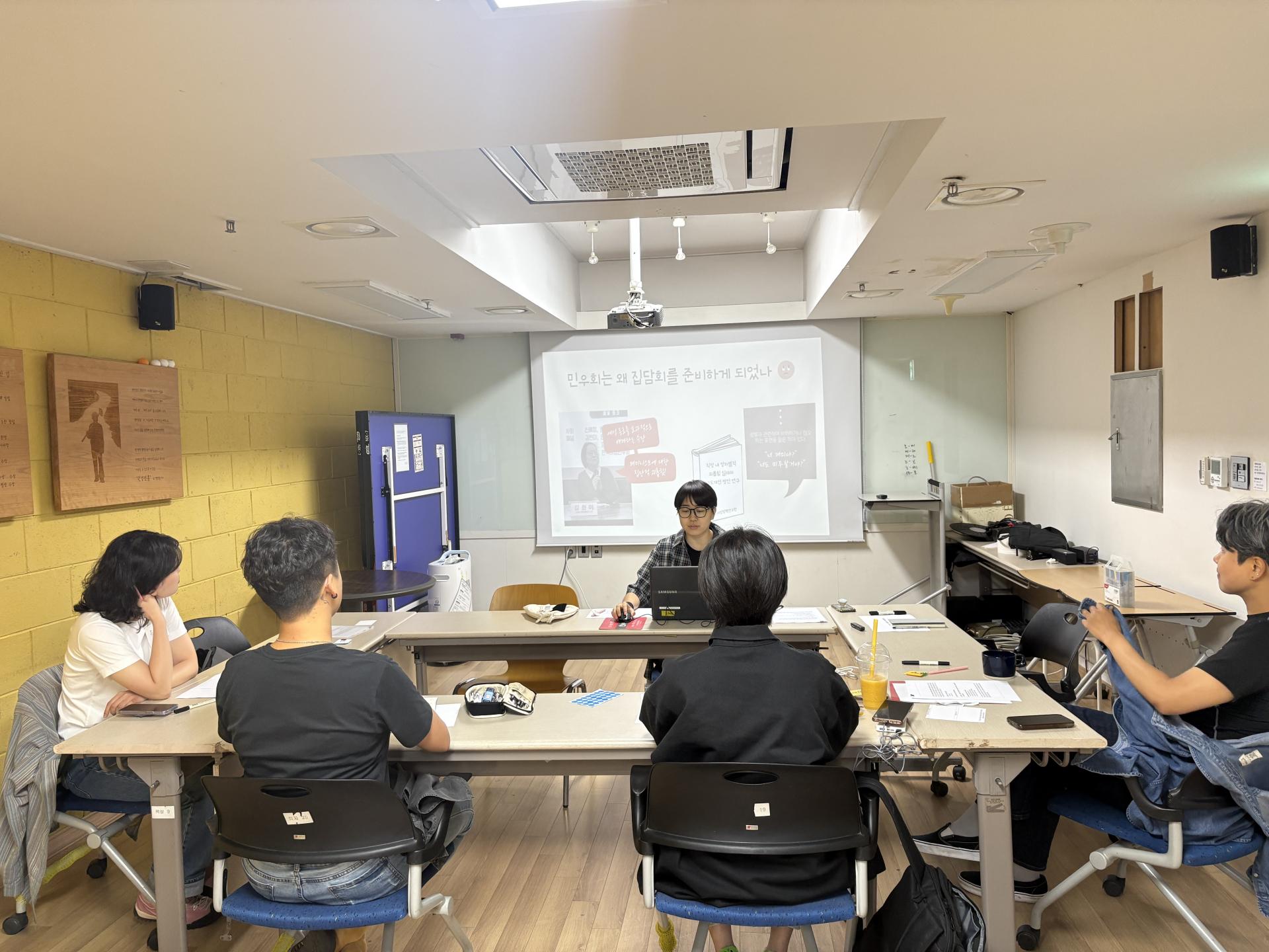 앞쪽에 빔프로젝터를 통해 집담회 진행 PPT가 보여지고 있다. 노트북을 앞둔 진행자를 중심으로 양쪽에 한 명, 그리고 맞은 편에 두 명의 참여자가 앉아서 화면에 집중하고 있는 모습이다.